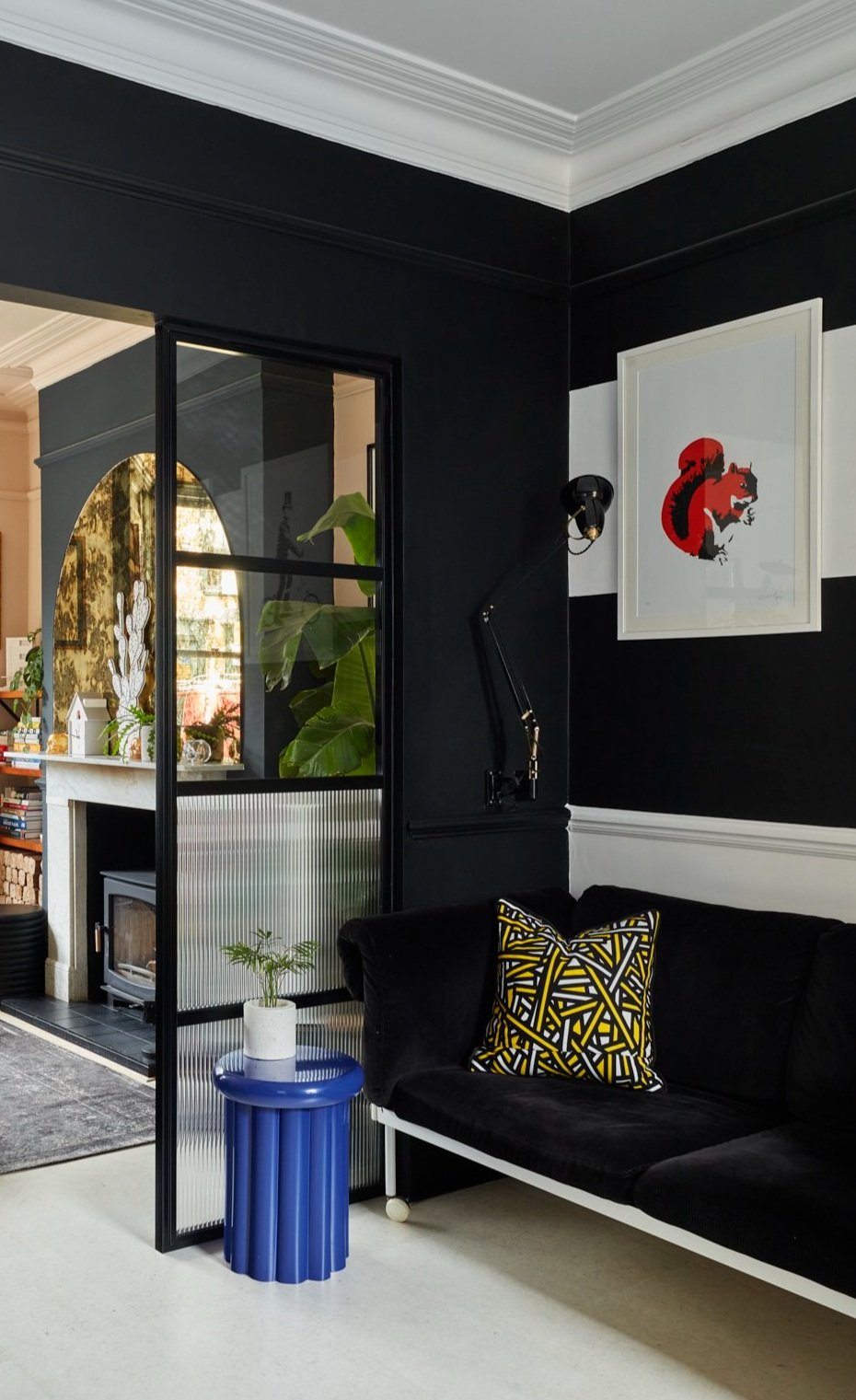 Modern living room with black walls, featuring a black sofa, a geometric-patterned cushion, and a blue side table with a small plant. A framed artwork of a red and black squirrel is on the wall. Glass door separates this room from another with plants and a fireplace.