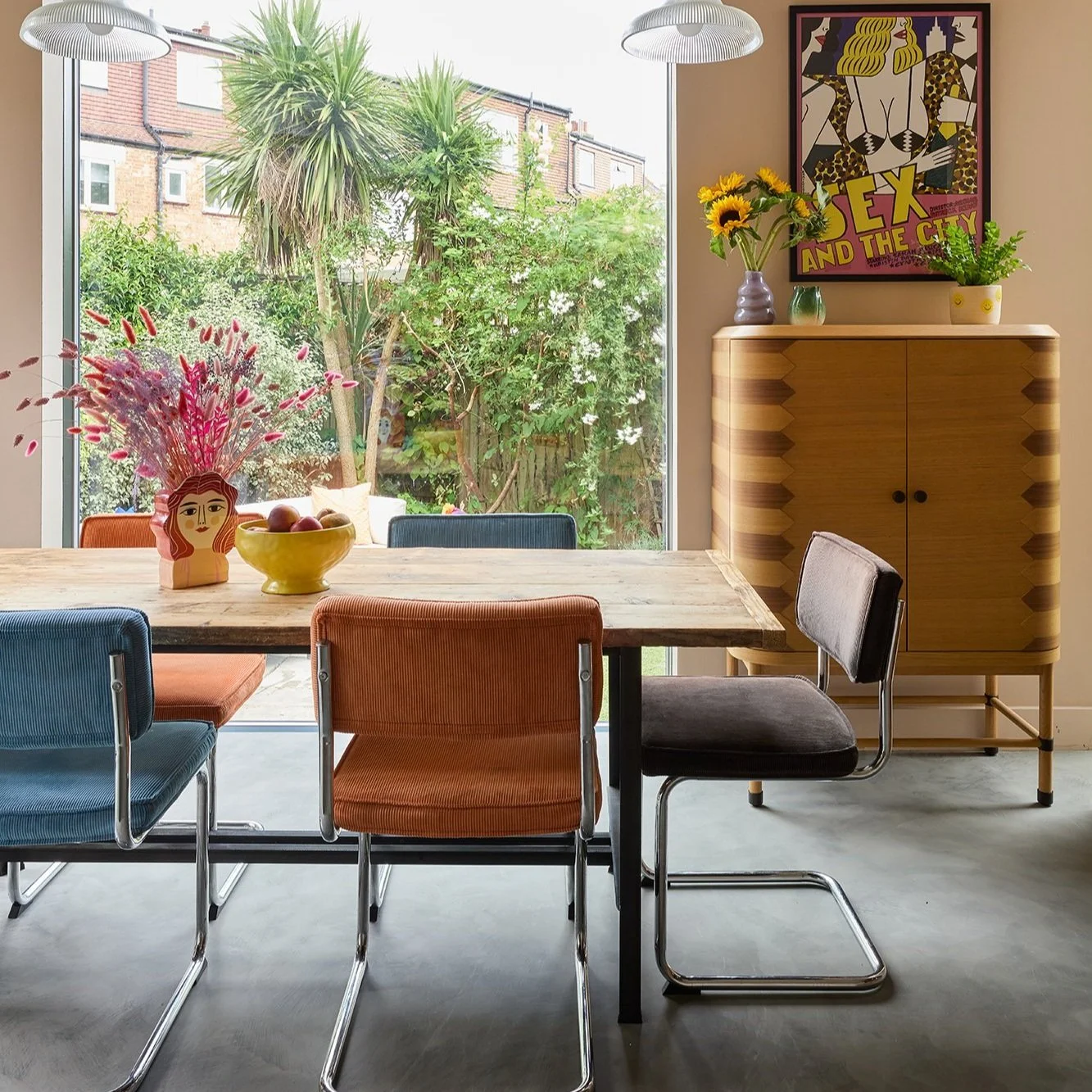 Dining room with colorful chairs around a wooden table, decorative plants, a cabinet, a poster on the wall, and a large window showing a garden view.