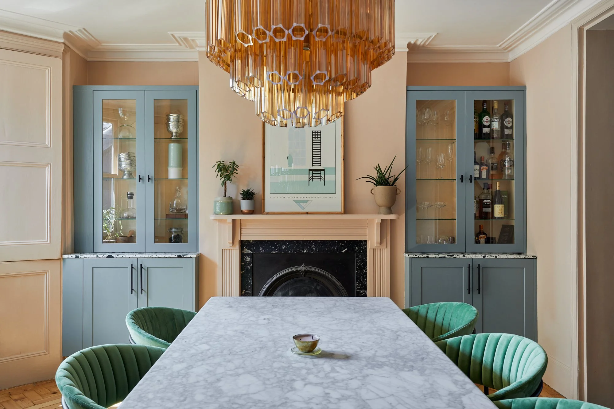 Dining room with marble table, green chairs, modern chandelier, two teal cabinets with glass doors, a fireplace with decorative items, and a framed artwork above.