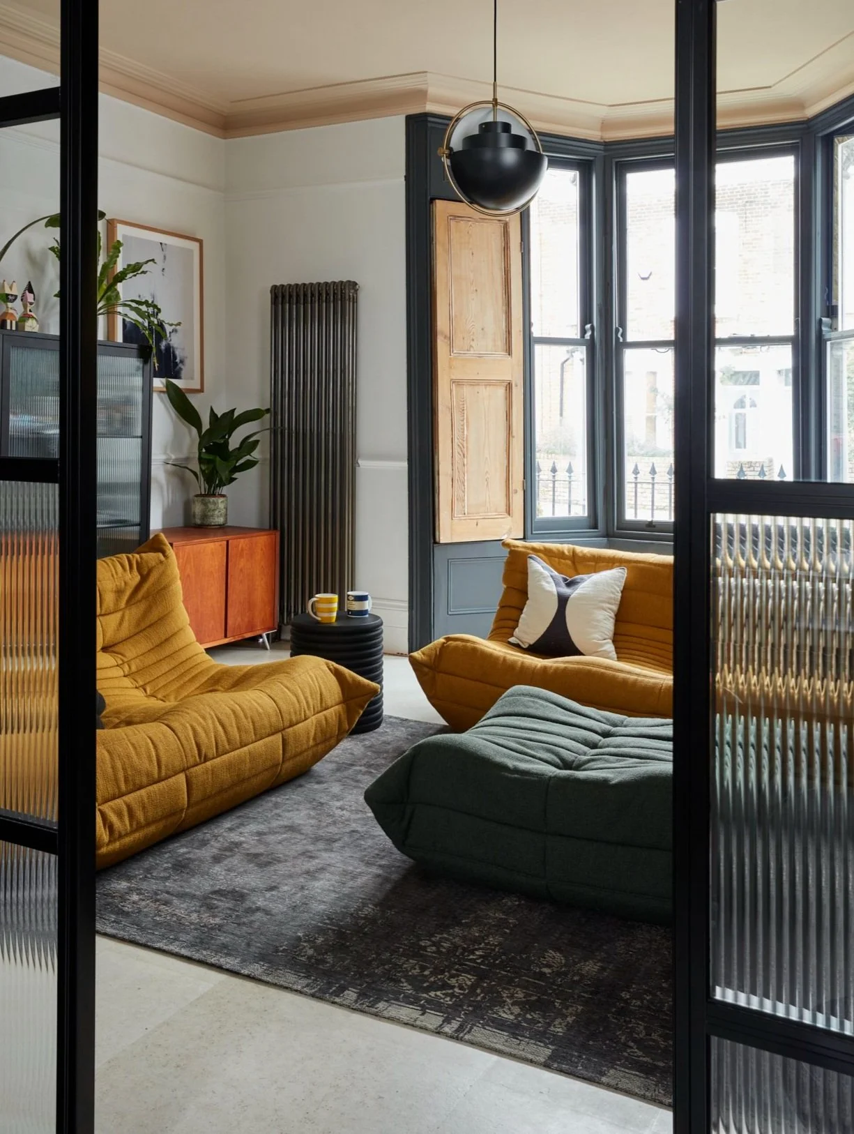 Modern living room with retro-style yellow and green couches, black pendant light, large windows, patterned rug, potted plant, and a wooden sideboard.