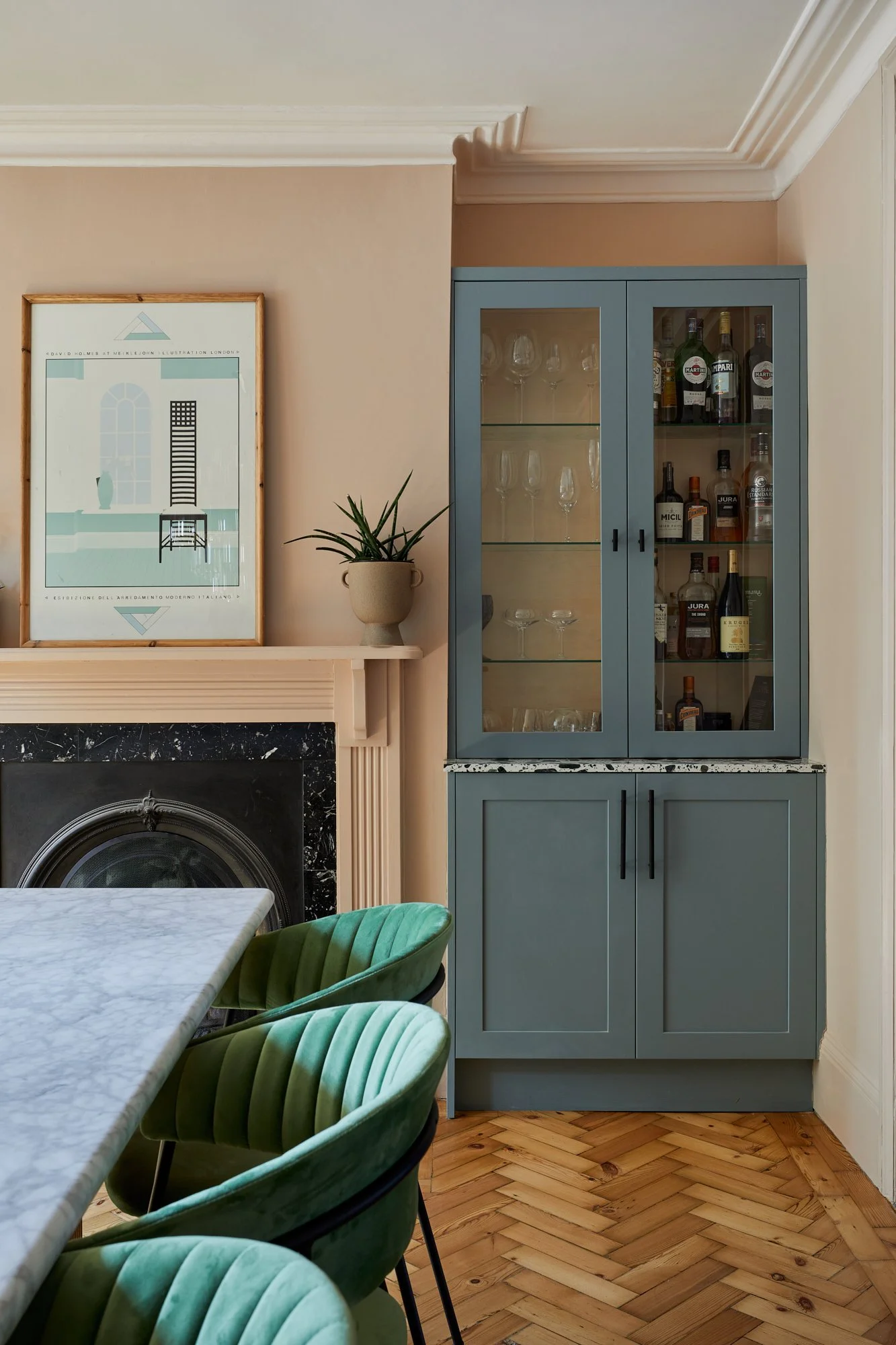 Interior dining area with blue cabinet, glassware, liquor bottles, green velvet chairs, marble table, framed wall art, plant on mantle, and herringbone wood floor.