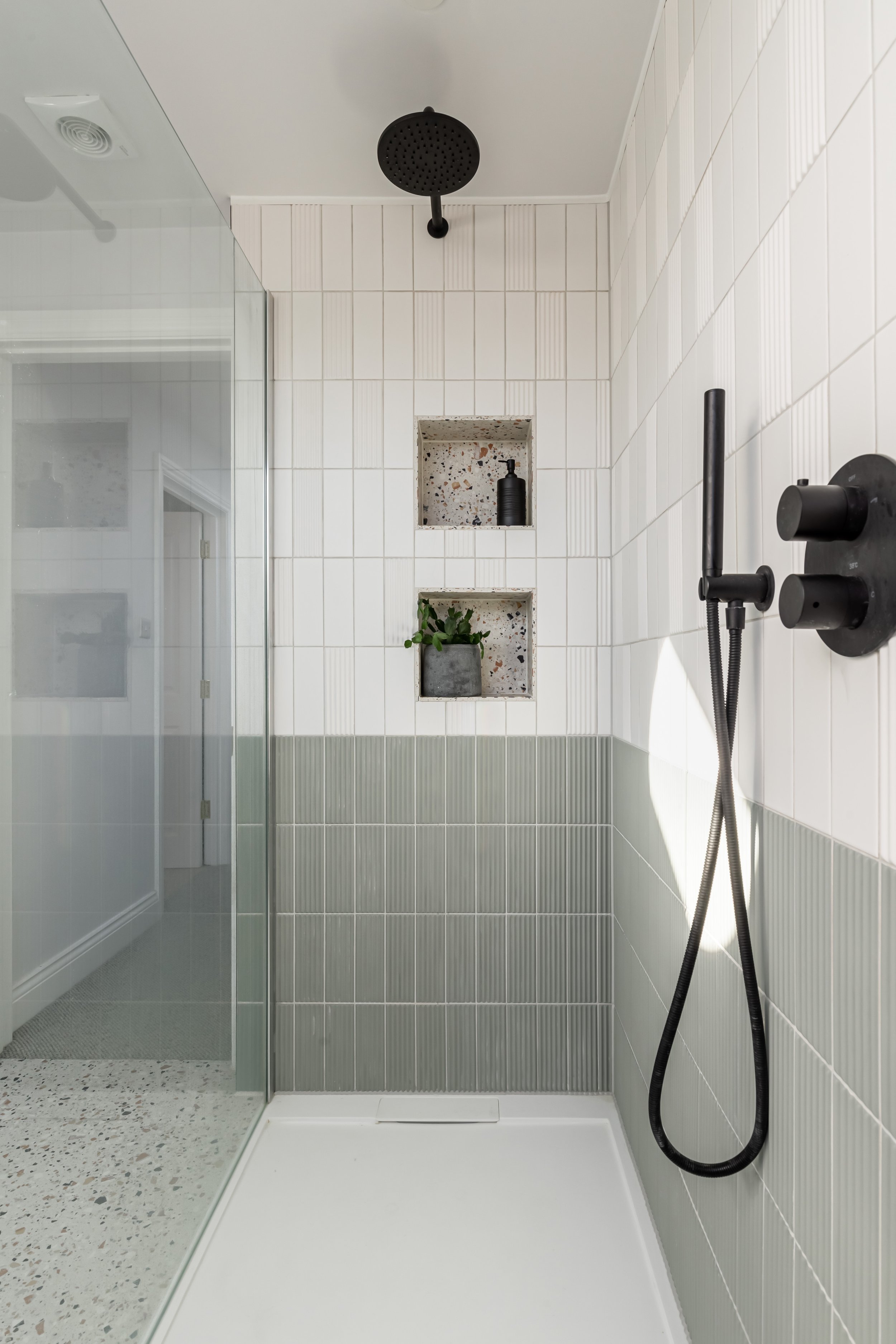 Modern bathroom shower area with overhead black showerhead, half-tiled walls in white and sage green, a black handheld shower, and two recessed shelves holding a plant and a black bottle.