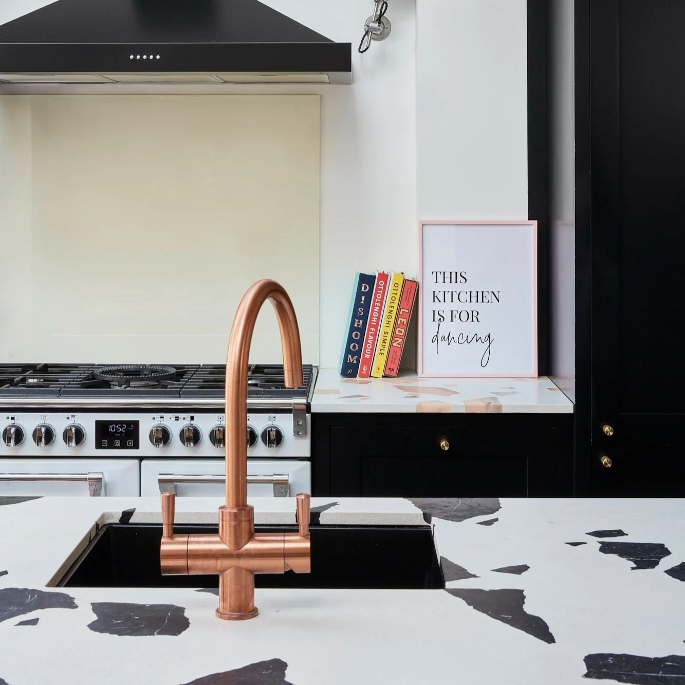 Modern kitchen with a copper faucet, black and white countertop, a stove with knobs, and a black range hood. A framed sign reads "This kitchen is for dancing," next to several cookbooks.