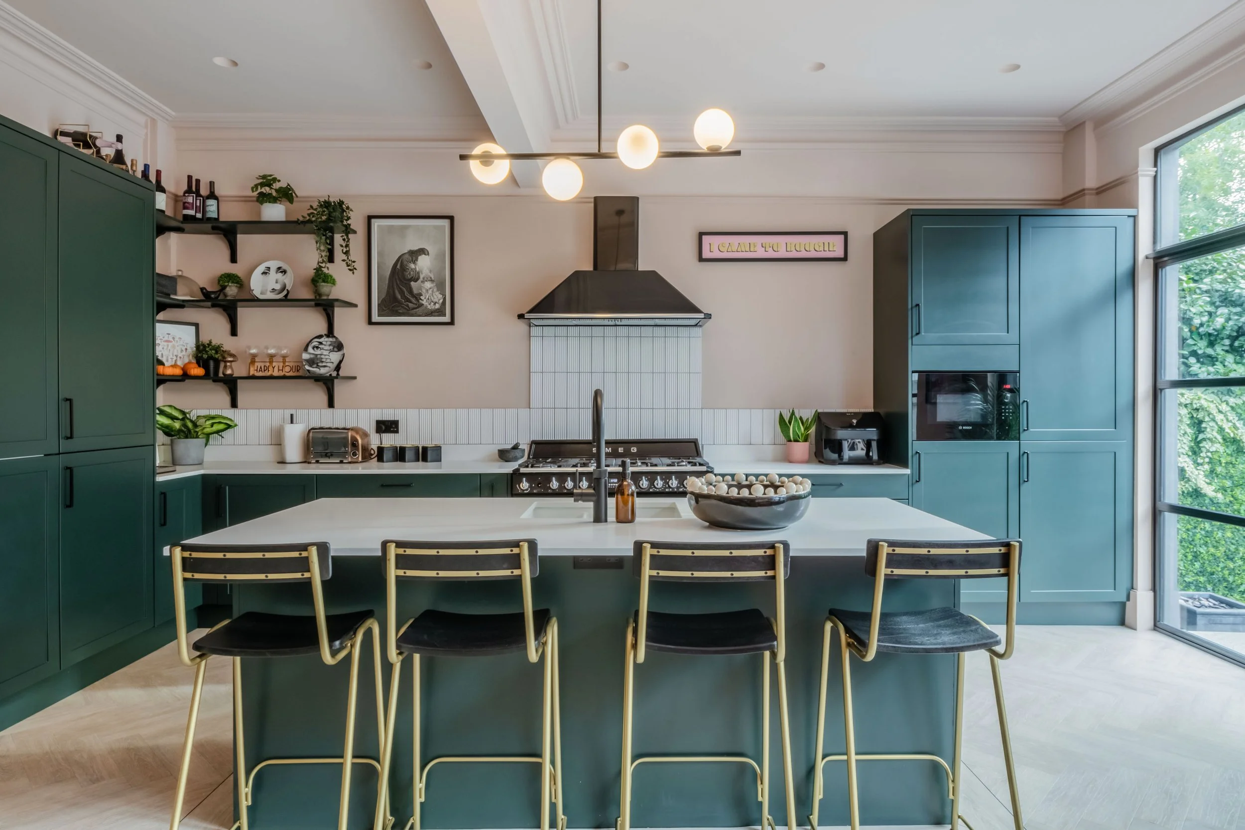 Modern kitchen with green cabinets, a central island with three bar stools, black range hood, open shelves with decor items, and a window offering natural light.