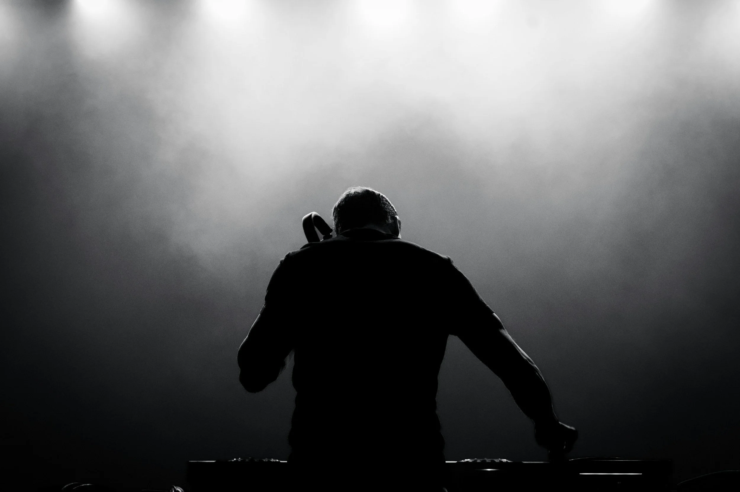 Black and white photo of a DJ performing on stage, with a silhouette of a person wearing headphones and standing behind DJ equipment, against a bright background.