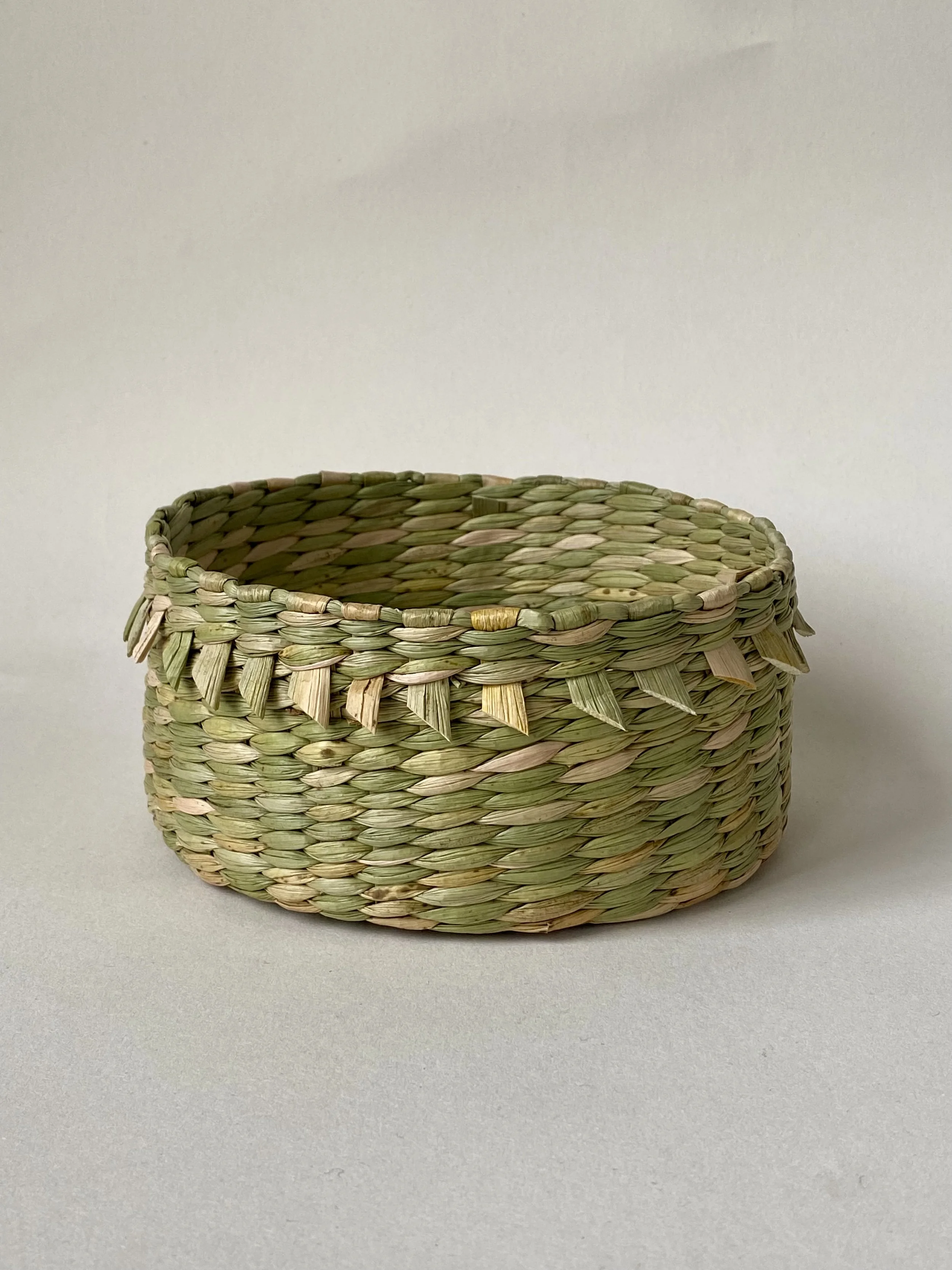 A cylindrical basket created in a pairing weave with a delicate tassel border. Ella Merriman finds the meditative state that she reaches whilst weaving extremely calming and grounding. She works in tandem with the rush stems and attempts to respect e