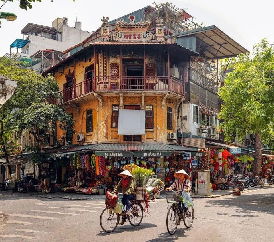 Two women riding bicycles with traditional conical hats in front of a colorful building with shops on the ground floor, trees, and motorcycles.