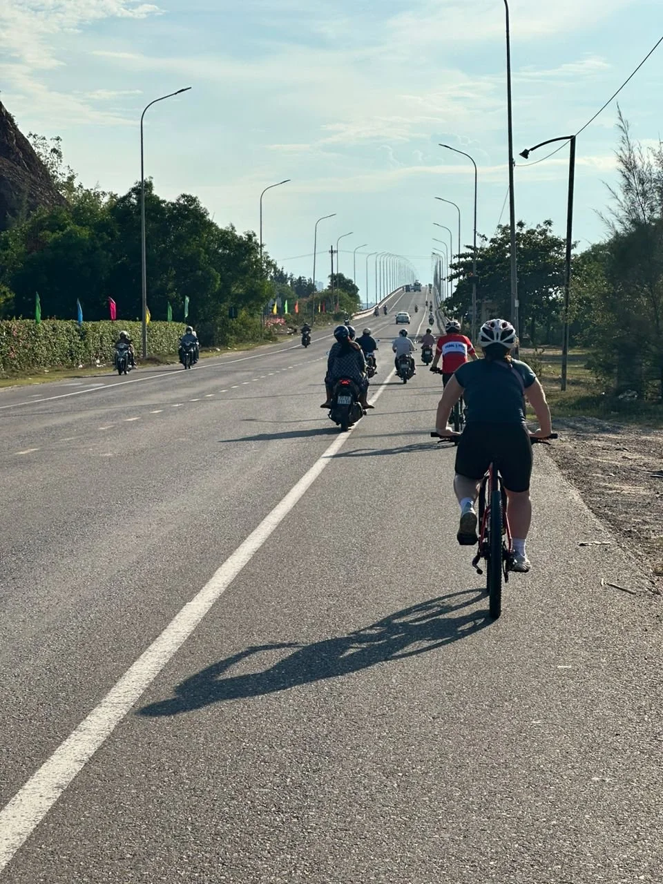 Cyclist riding on a paved shoulder next to a busy road with cars and motorcycles, green trees lining the sides, and a cloudy sky overhead.