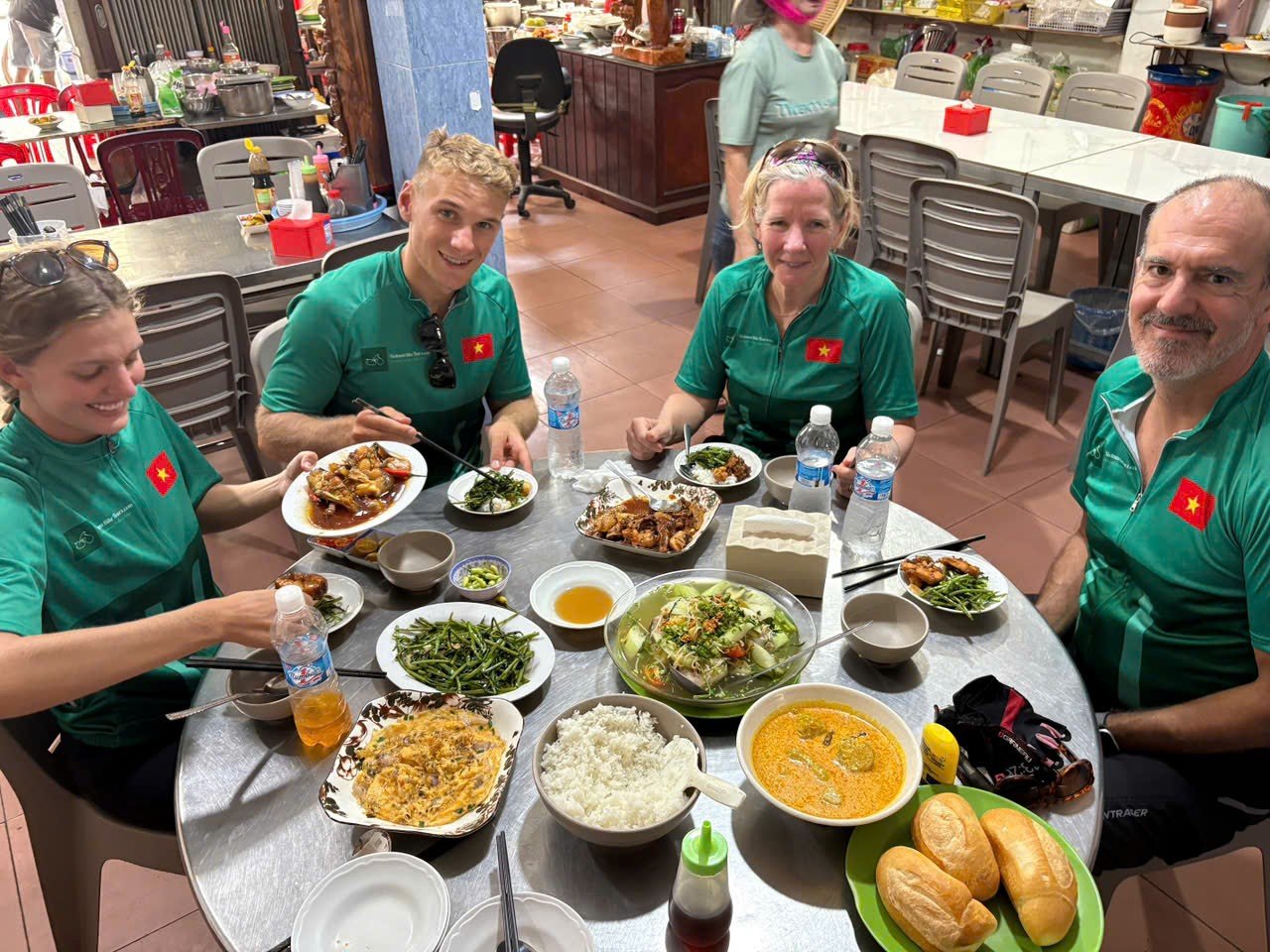 A group of four people wearing matching teal shirts with Vietnamese flags, enjoying a meal at a restaurant table filled with dishes, including rice, soup, vegetables, bread rolls, and drinks. They are smiling and appear to be in a casual dining environment with other tables and utensils visible in the background.