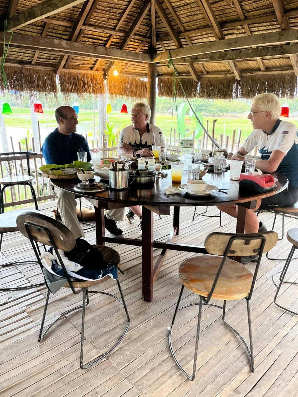 Three men seated around a round wooden table with various dishes and drinks inside a rustic, open-air pavilion with bamboo roof, overlooking a green landscape.