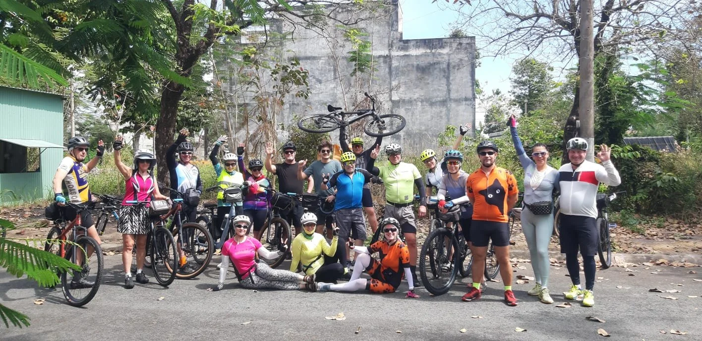 Group of cyclists with helmets and sportswear, posing with their bicycles on a city street during daytime, some standing and some sitting or lying on the ground, with a bicycle lifted in the air, trees, and a building in the background.