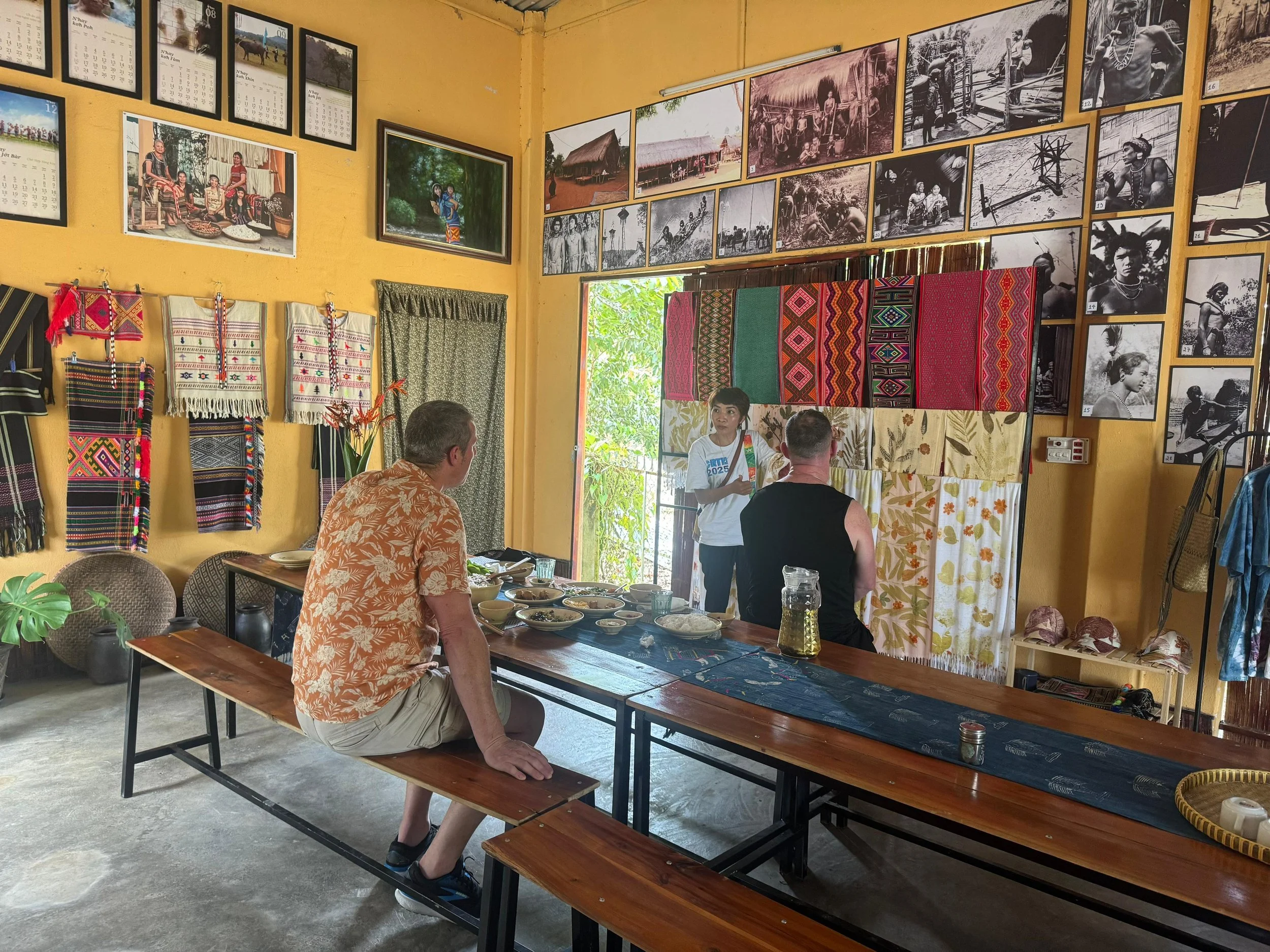 A room decorated with photos of indigenous people and scenes on yellow walls, with woven textiles hanging on display. Several people are sitting at a wooden table with food, and a woman is standing, speaking to them.