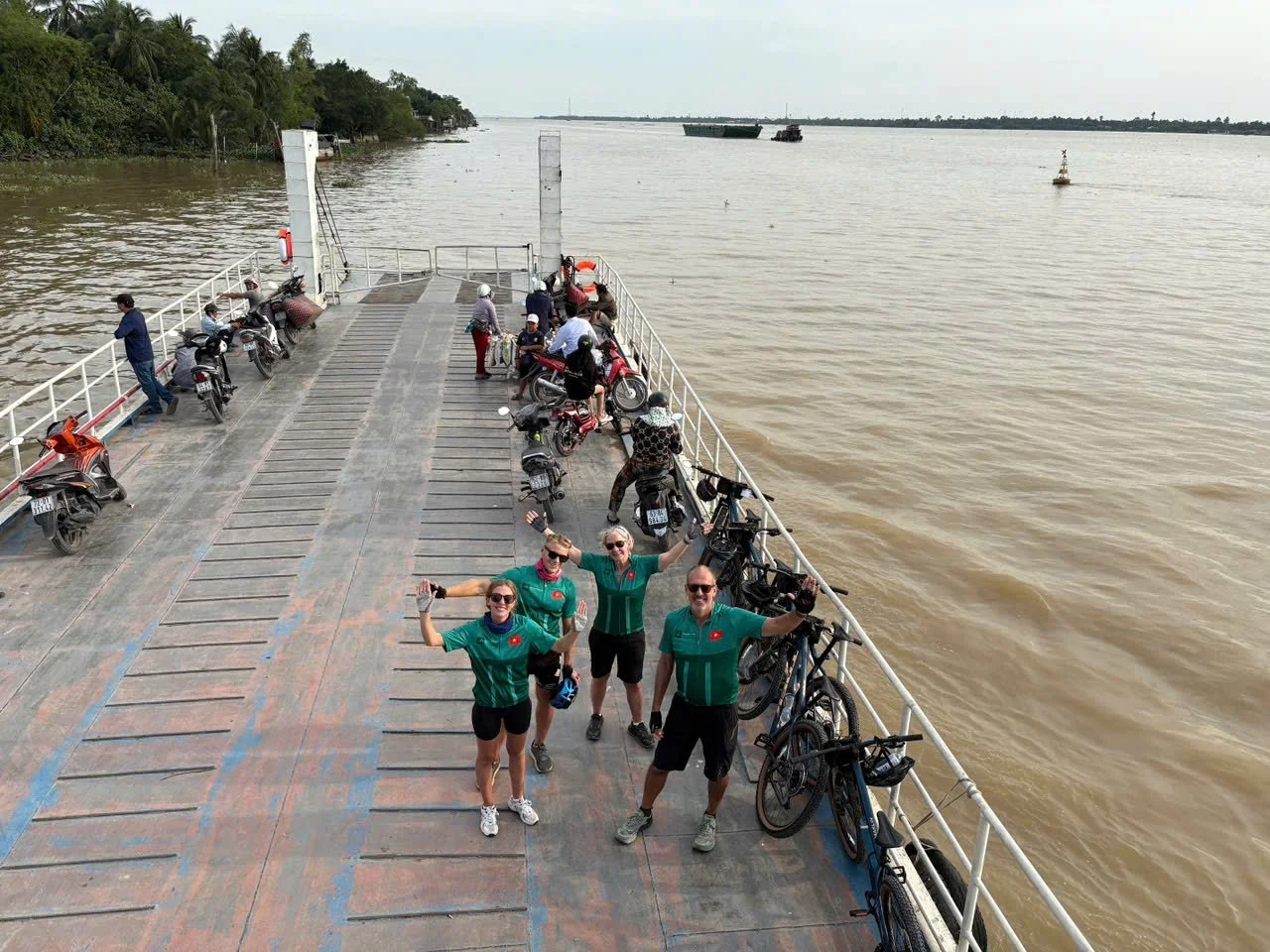 Group of four cyclists standing on a ferry boat with their bikes, smiling and posing for the photo, over a river with trees on the left bank and boats in the water.