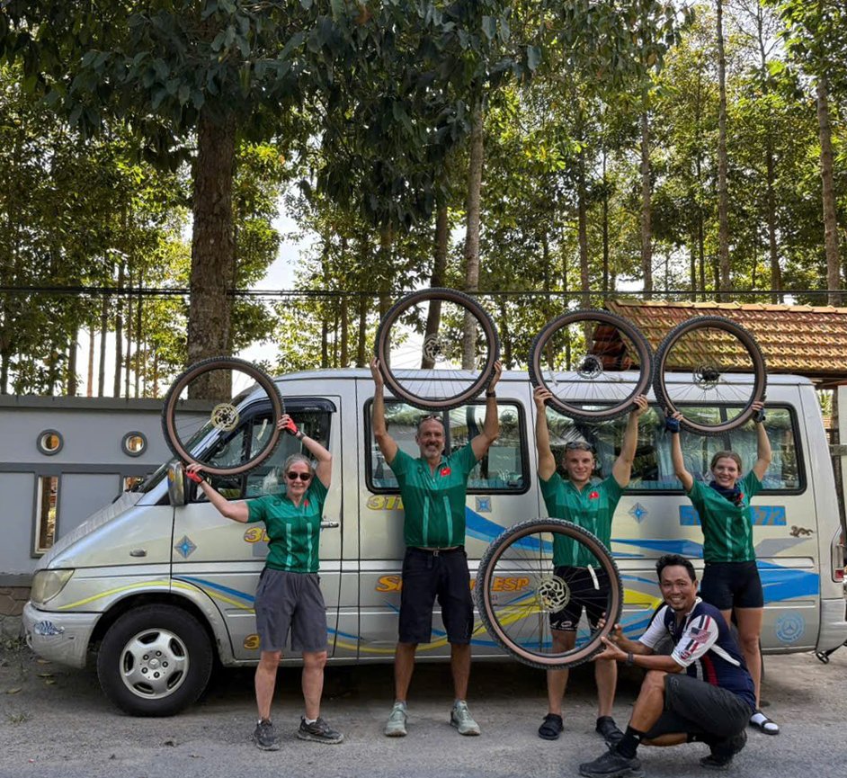 Five people, four standing and one kneeling, holding bicycle wheels in front of a van, outdoors with trees in the background.