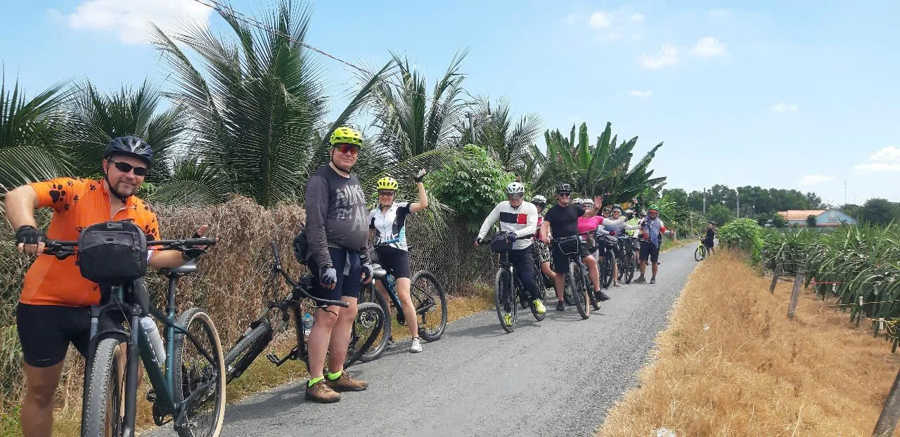 Group of cyclists on a rural road surrounded by tropical plants and trees in sunny weather.
