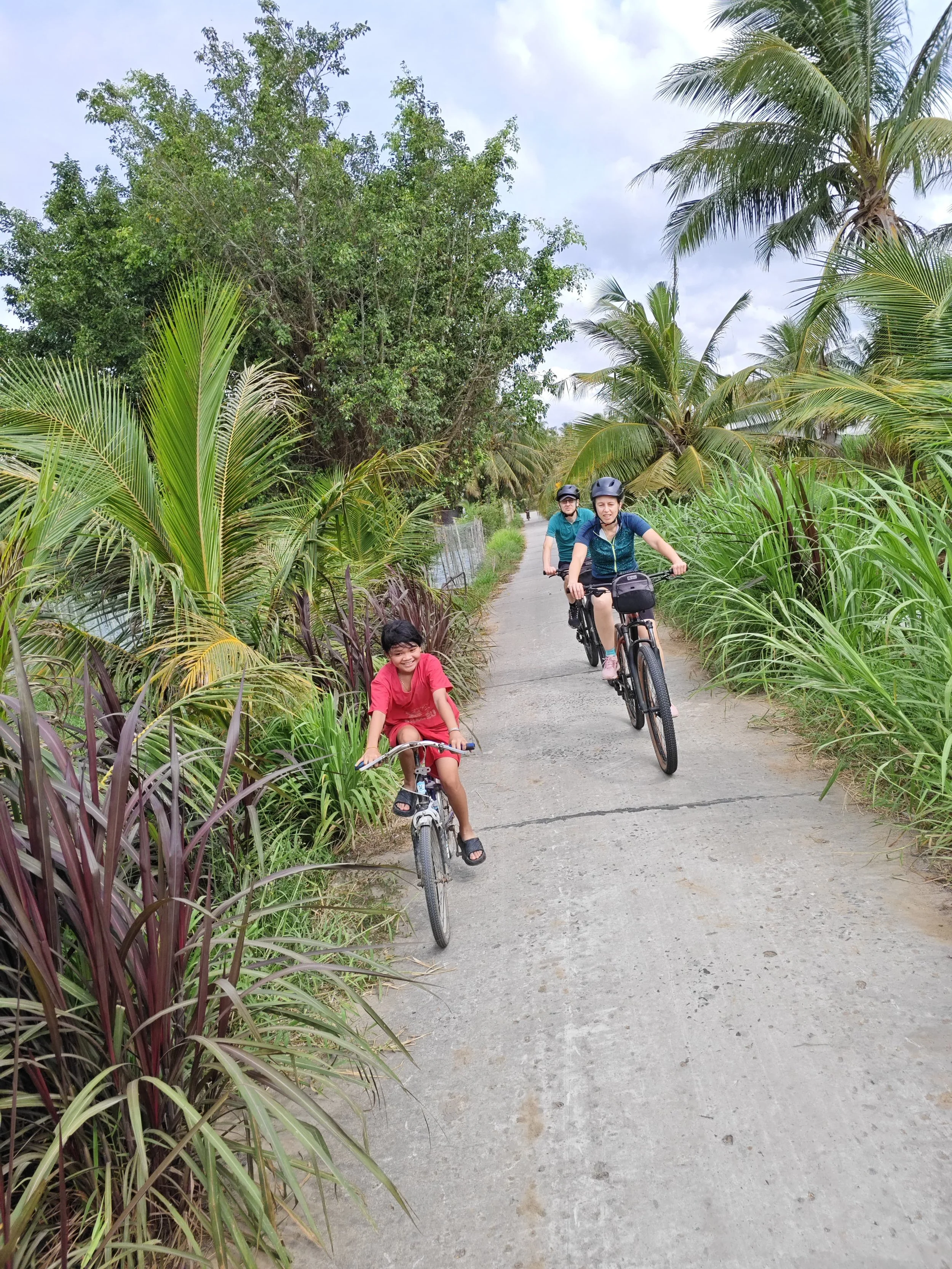 Two children riding bicycles on a narrow concrete path surrounded by tall green palm trees and shrubs, under a cloudy sky.