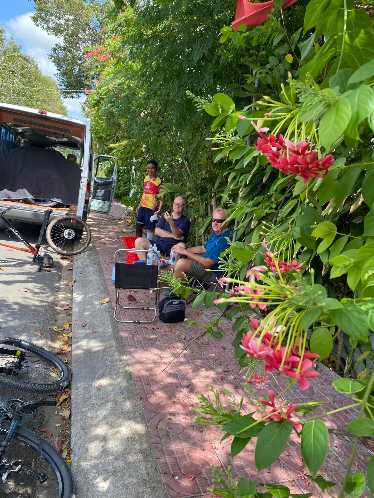 Three men sitting and standing by a sidewalk, with bicycles and a van nearby, surrounded by green trees and pink flowers, on a sunny day.