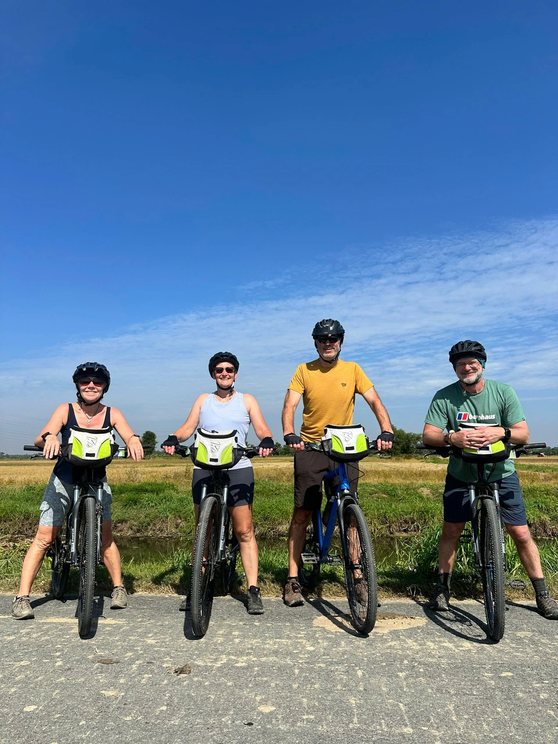 Five people standing with bikes outdoors on a sunny day, all wearing helmets and casual clothes, smiling at the camera.