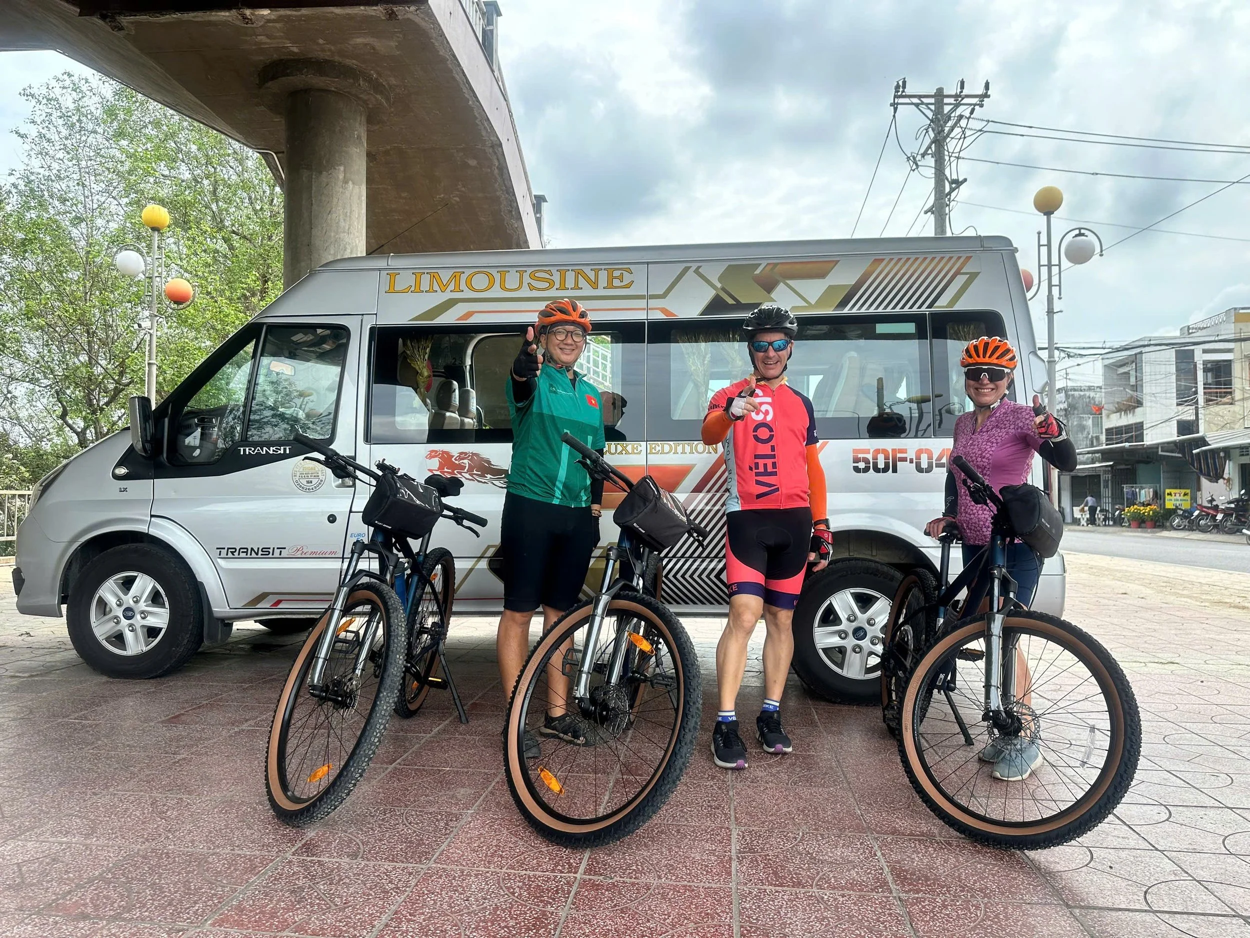 Three cyclists smiling and posing with thumbs up in front of a silver van labeled 'Limousine' parked on a brick-paved street, with trees, electric poles, and a building in the background.