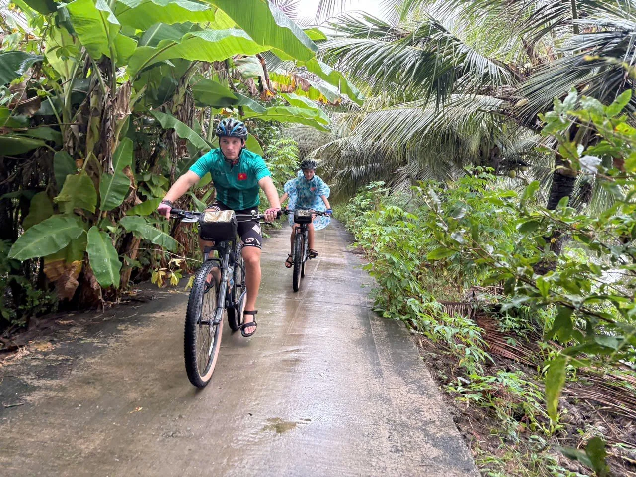 Two children riding mountain bikes on a narrow concrete path surrounded by lush tropical green plants and banana trees.