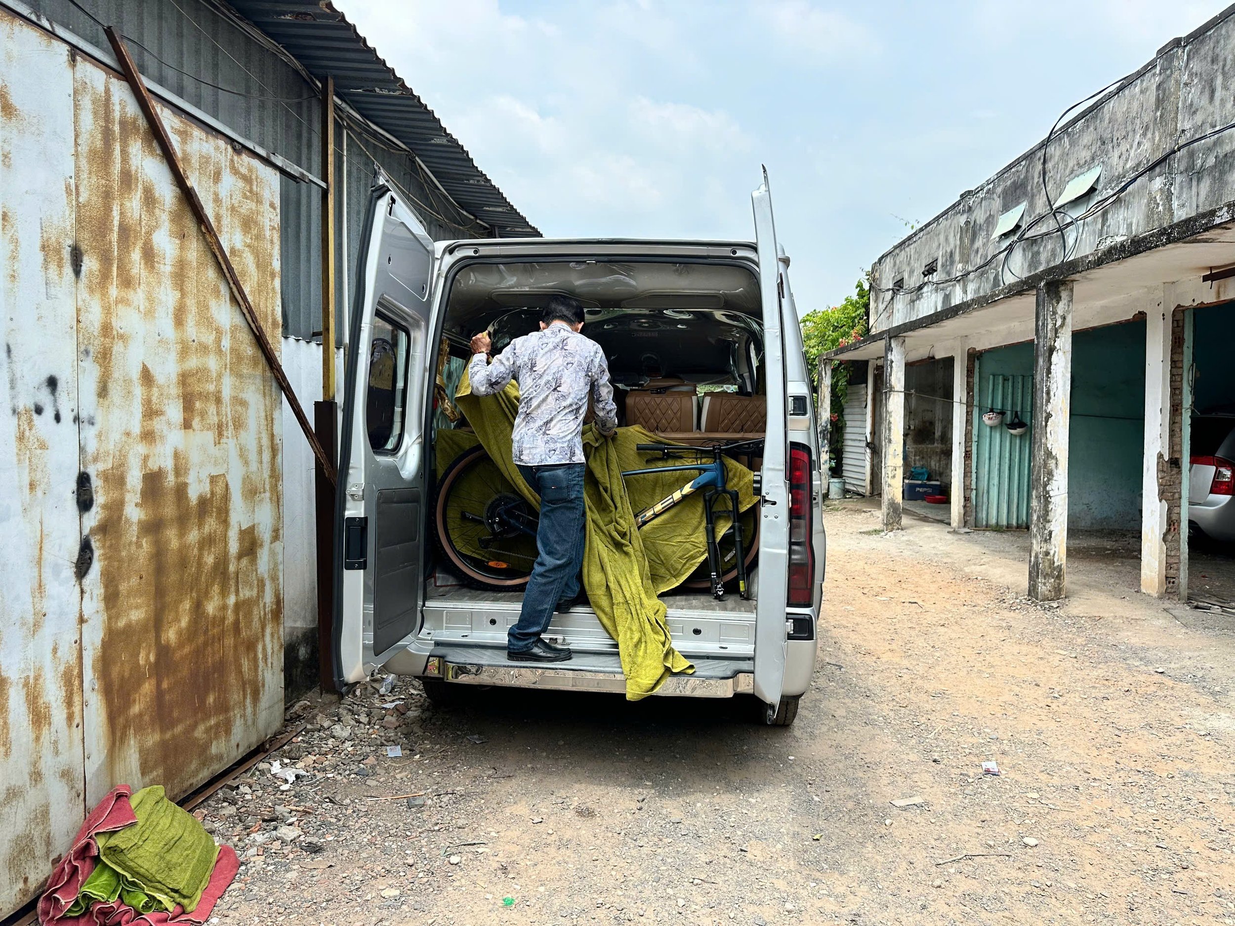 A man loading yellow and green blankets into the back of a silver van parked between weathered buildings in a rustic area.