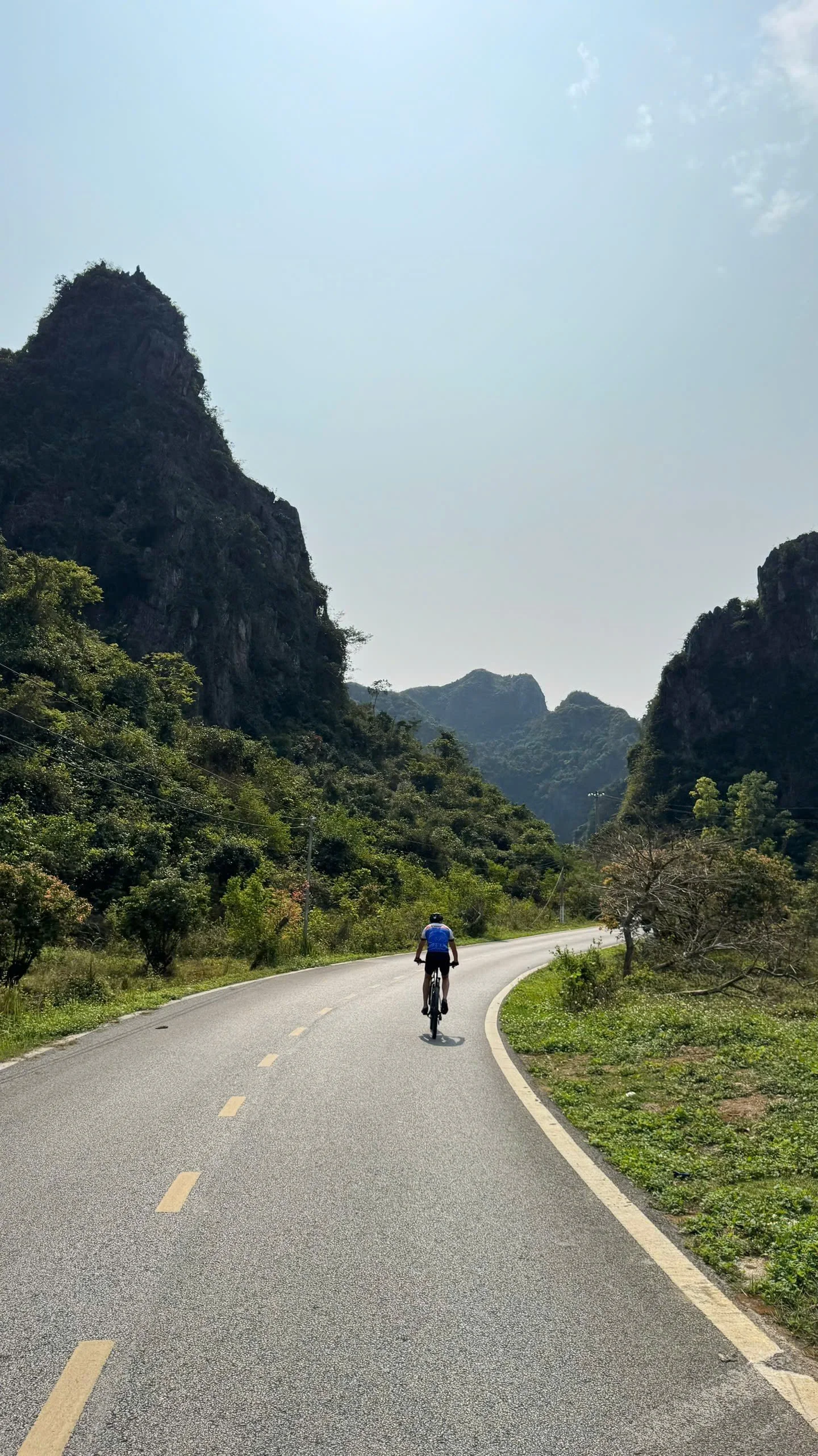 A person riding a bicycle on a winding mountain road surrounded by green vegetation and tall rocky hills under a clear sky.