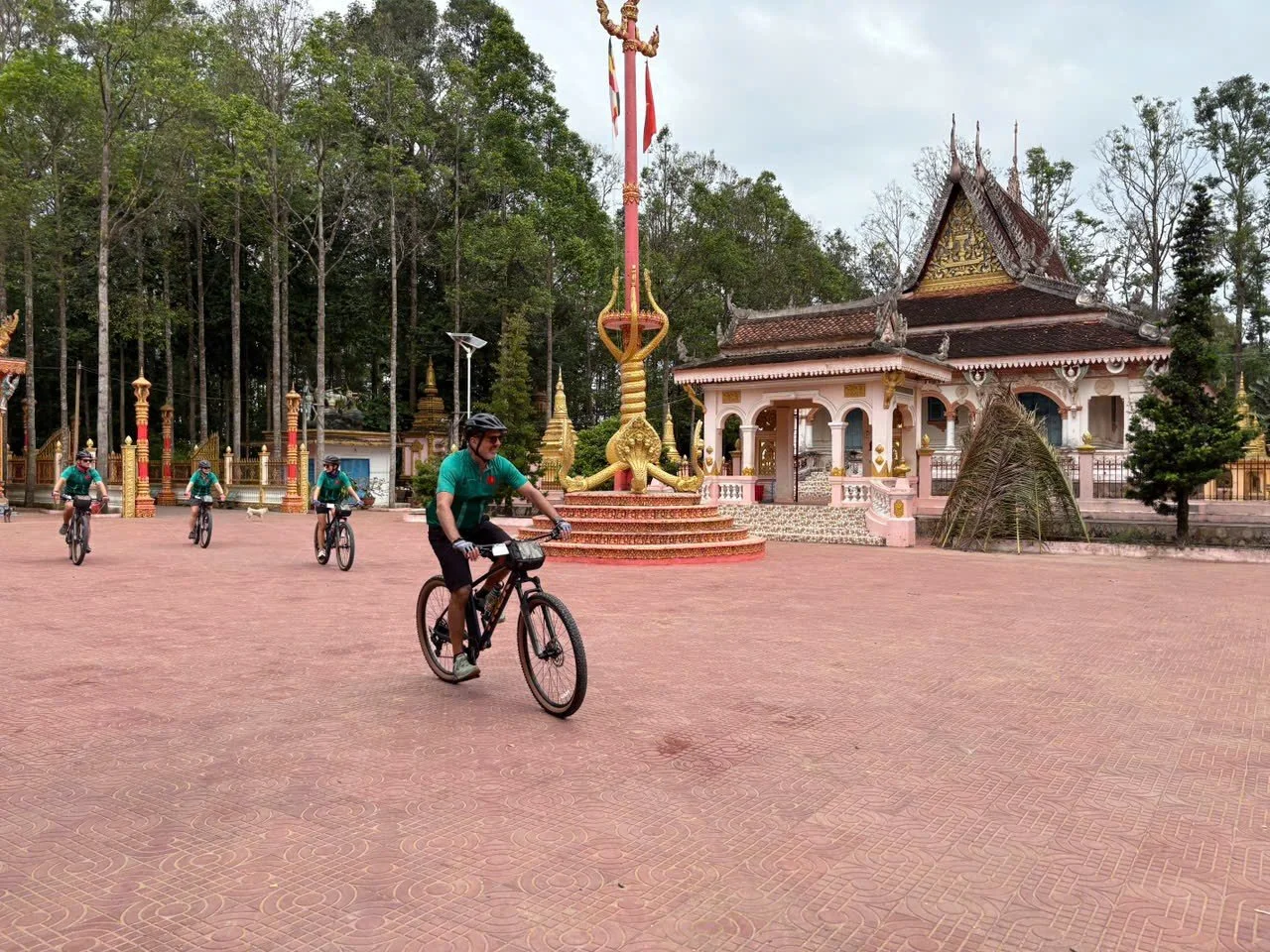 Group of people riding bicycles on a paved area near traditional Asian-style buildings and lush green trees.