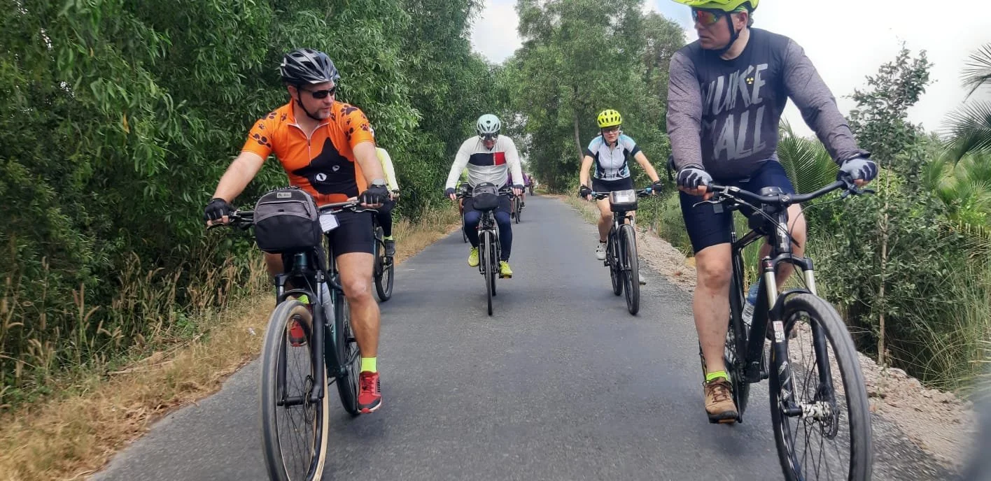 Four people cycling on a paved road surrounded by lush green trees and bushes, with two men in the foreground and two women behind them, all wearing helmets and athletic clothing.