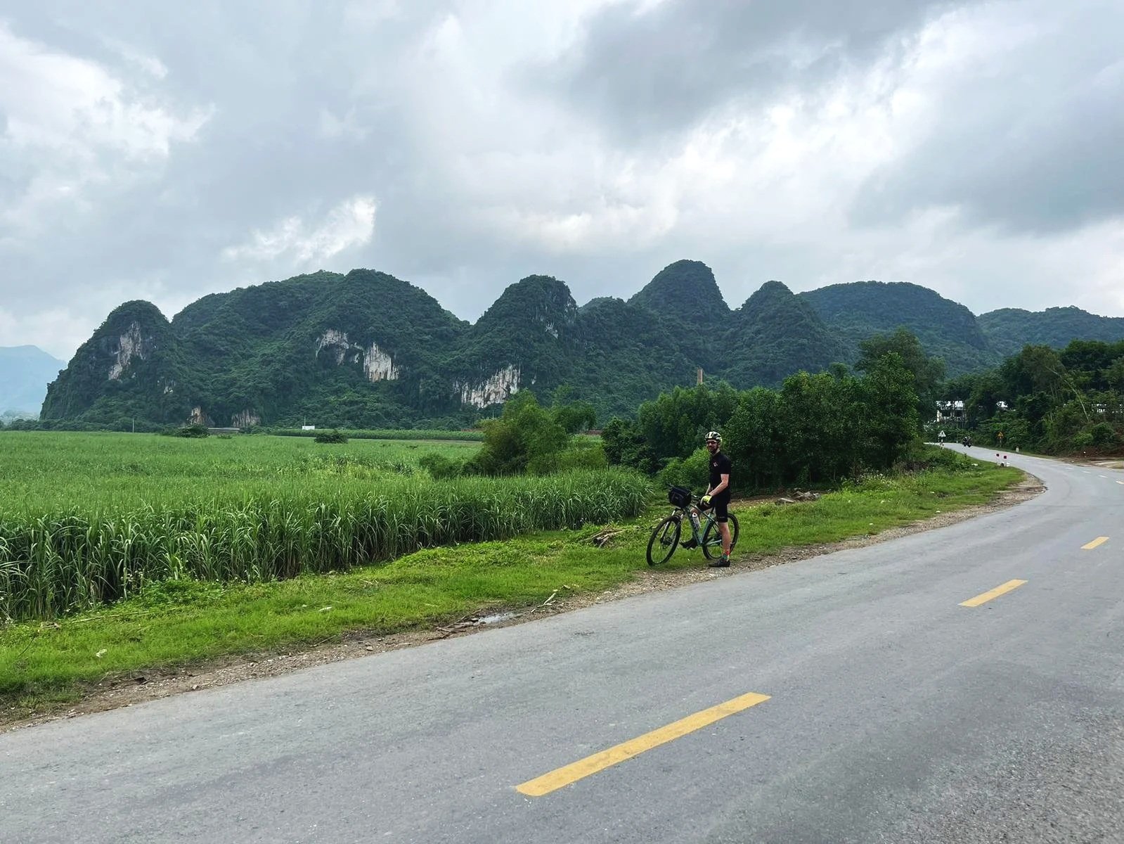 A person standing with a bicycle on the side of a winding road, lush green fields in the background, and mountains with dense forest under a cloudy sky.