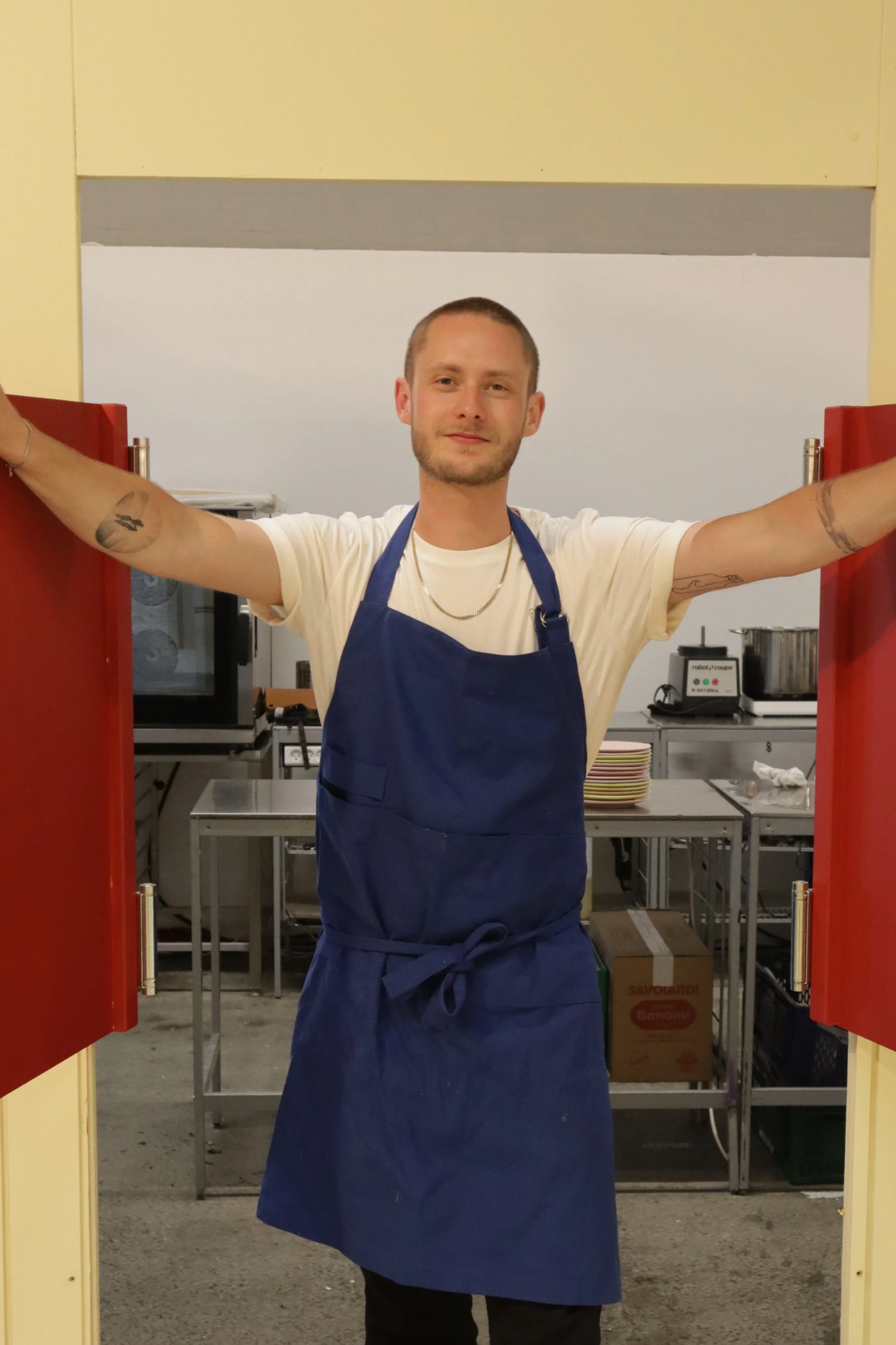A young man wearing a white t-shirt, blue apron, and gold chain, standing in a kitchen, holding open red cabinet doors with arms outstretched.