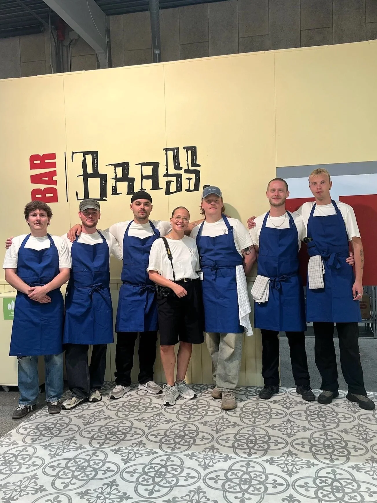 En gruppe på syv personer står foran en gul væg med et skilt, hvor der står “Bar” og “Barss”. Alle har hvide skjorter og blå forklæder på, og nogle bærer også kasketter eller briller.