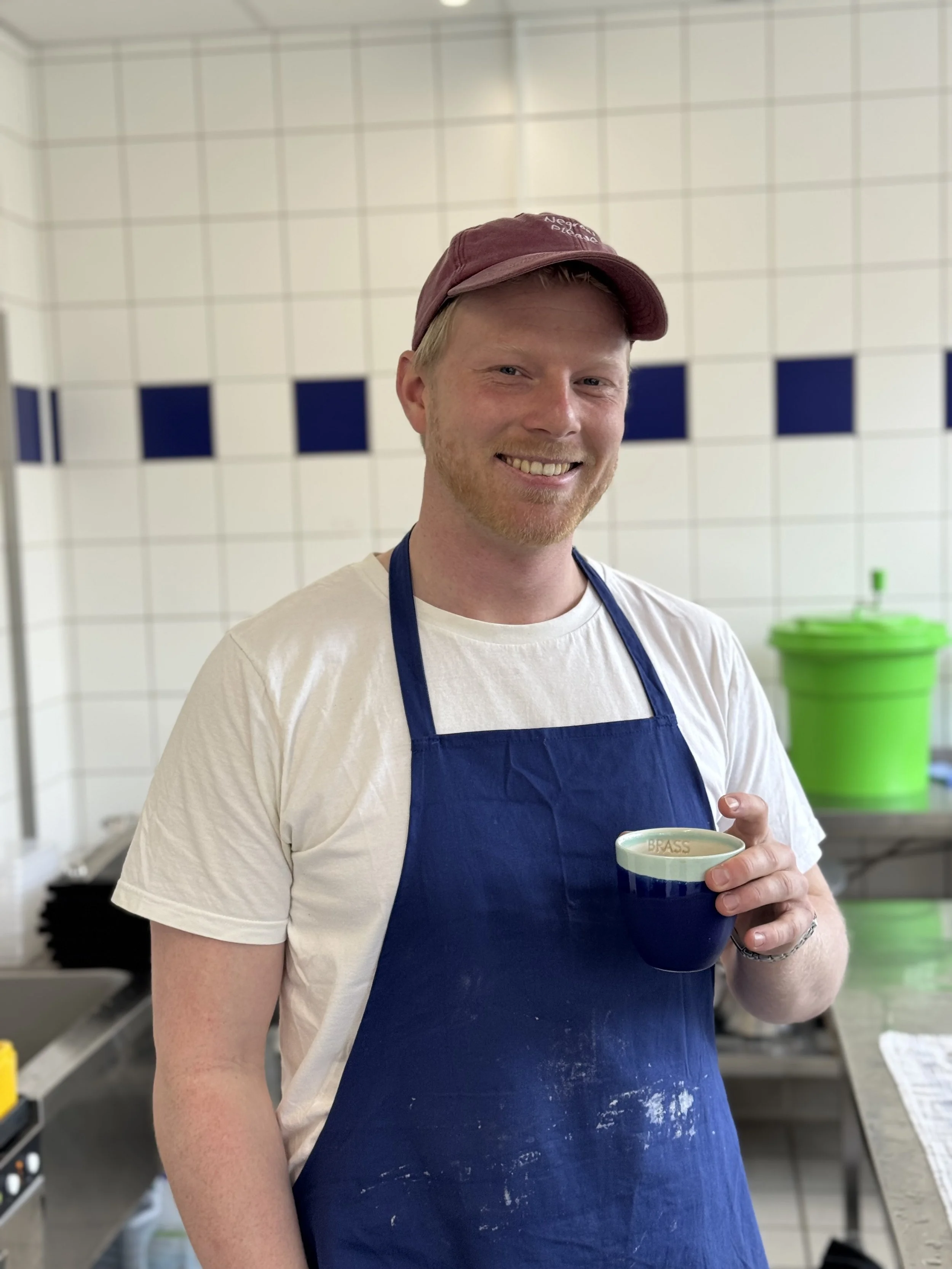 Smiling young man in a kitchen, wearing a maroon cap, white t-shirt, and a blue apron, holding a dark blue mug.