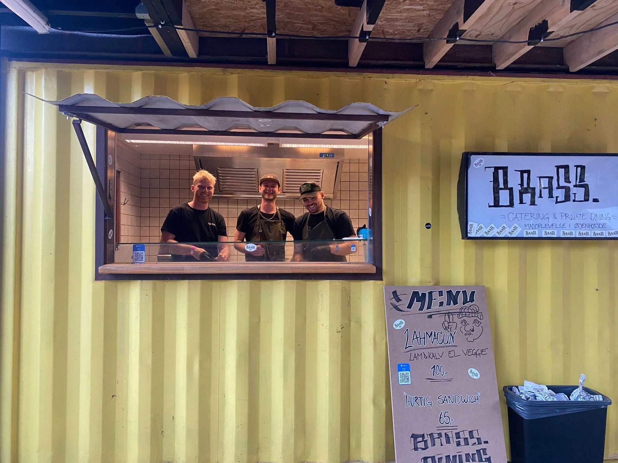 Tre smilene kokke i et serverings vindue til en festival. Et stort skilt der står BRASS på og en menu med en masse lækre madvare.