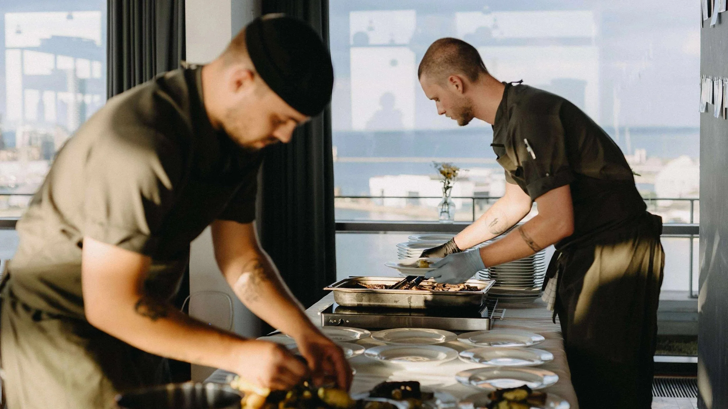 To kokke portionsanretter mad ved et bord, til et bryllup, i et lyst spiseområde med store vinduer og udsigt til udendørsområdet.