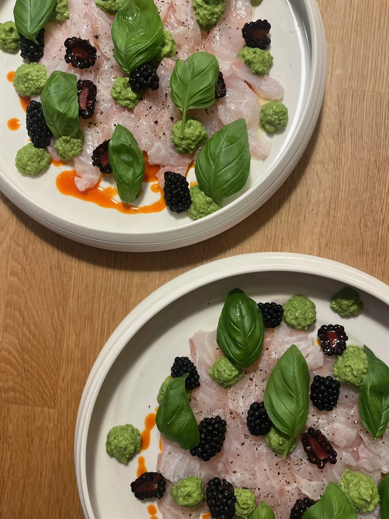 To tallerkener med flot ceviche toppet med basilikum, brombær, ærtepure og olie.