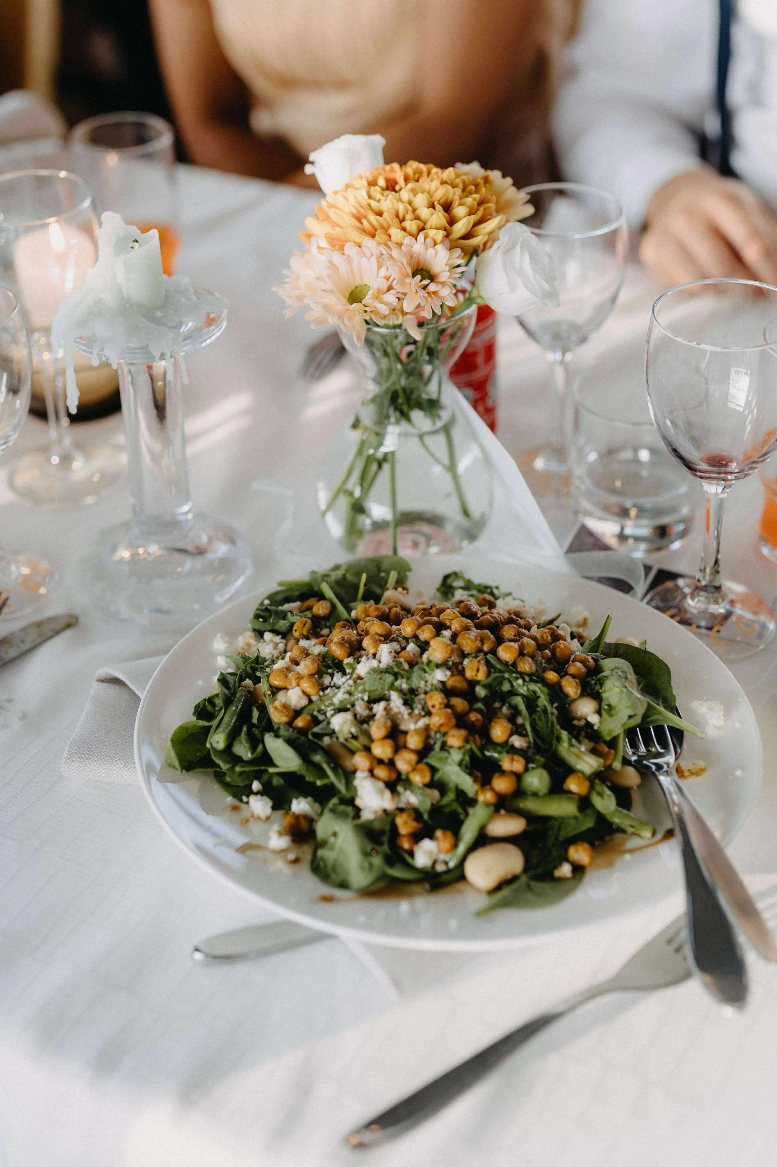 En tallerken med blandet grøn salat toppet med kikærter, fetaost og urter på en hvid dug, med en glasvase med lyserøde og orange blomster og tomme glas i baggrunden.