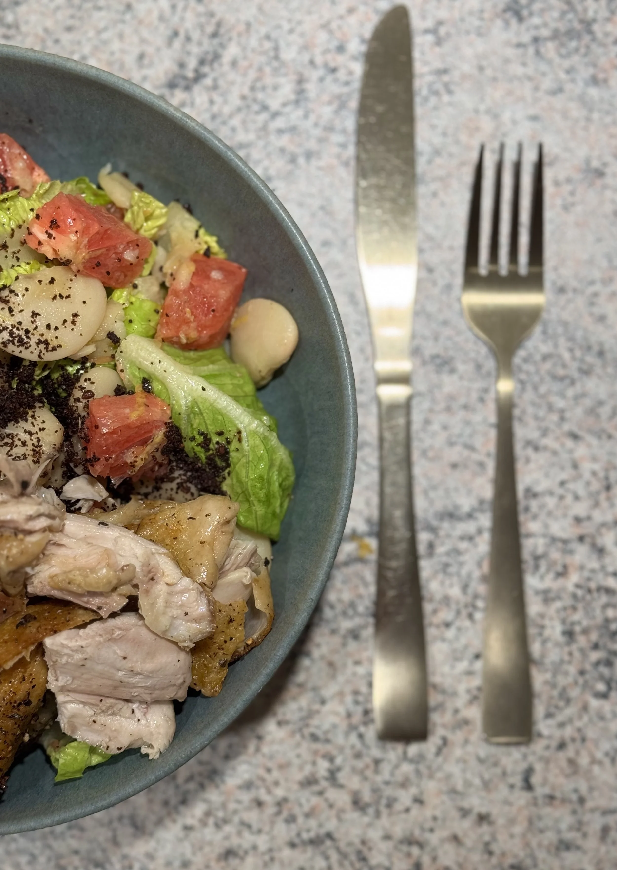 En salat med salatblade, bønner, kylling, tomater og sort peber i en blå skål på en plettet bordplade, med en gaffel og en kniv ved siden af.