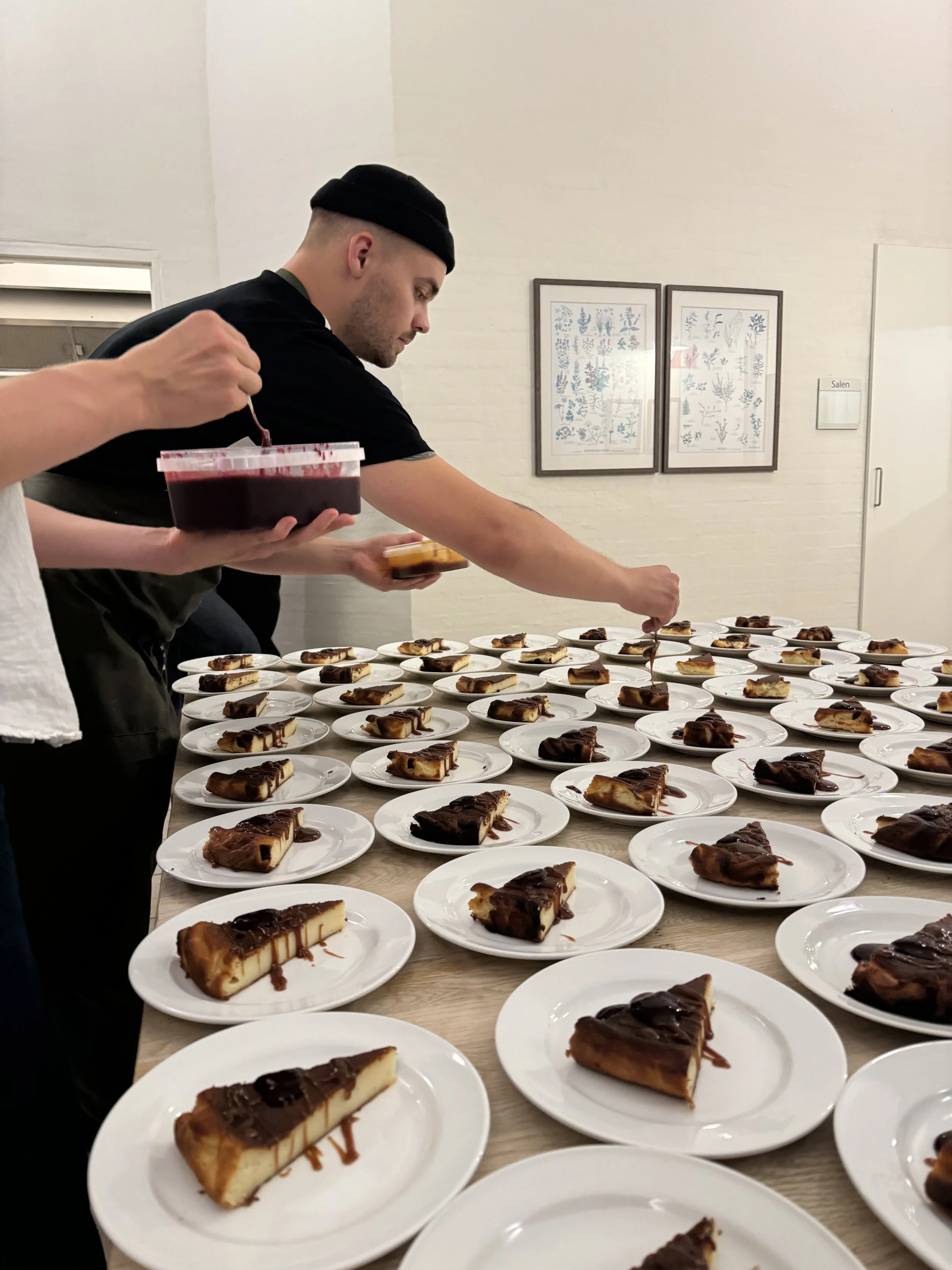 Mange tallerkener med Basque-cheesecake med bær kompot sat op på et stort bord, med en kok der anretter i baggrunden. 