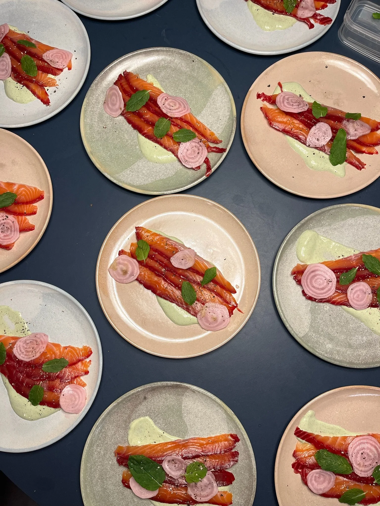 Flere tallerkener med graved laks, lyserøde radiseskiver, friske mynteblade og en grøn sauce på et mørkt bord.