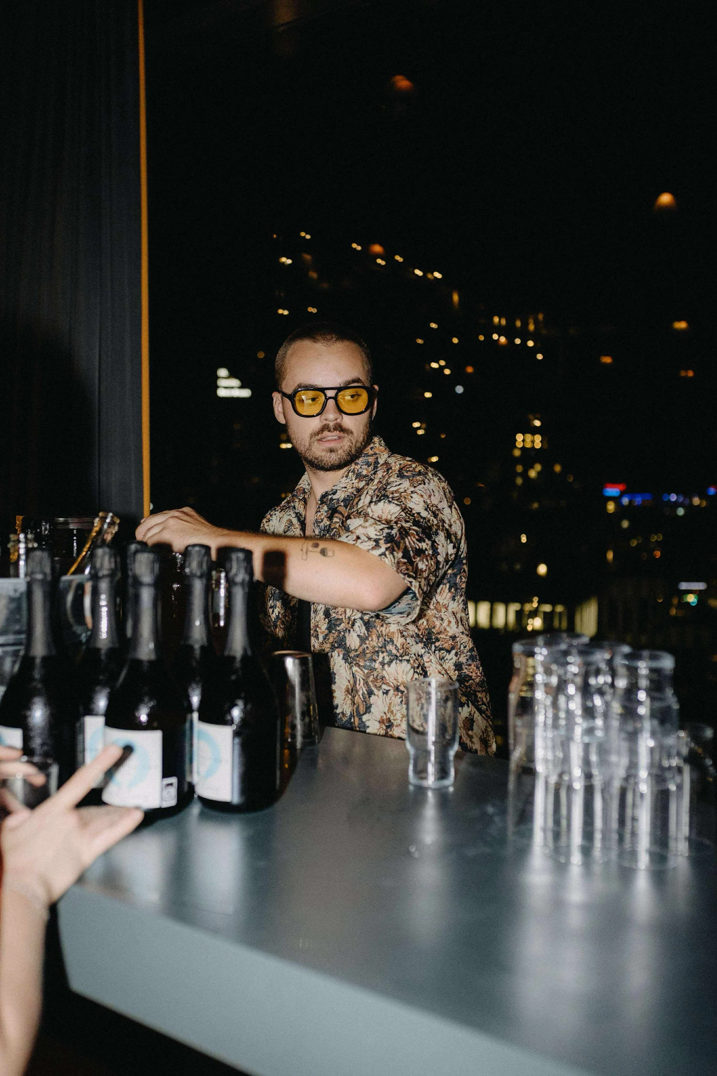 En bartender med gule biller og en blomstret skjorte bag en bar om aftenen, med en skyline i baggrunden.A man wearing yellow-tinted glasses and a floral shirt behind a bar at night, with a city skyline visible through the window in the background.