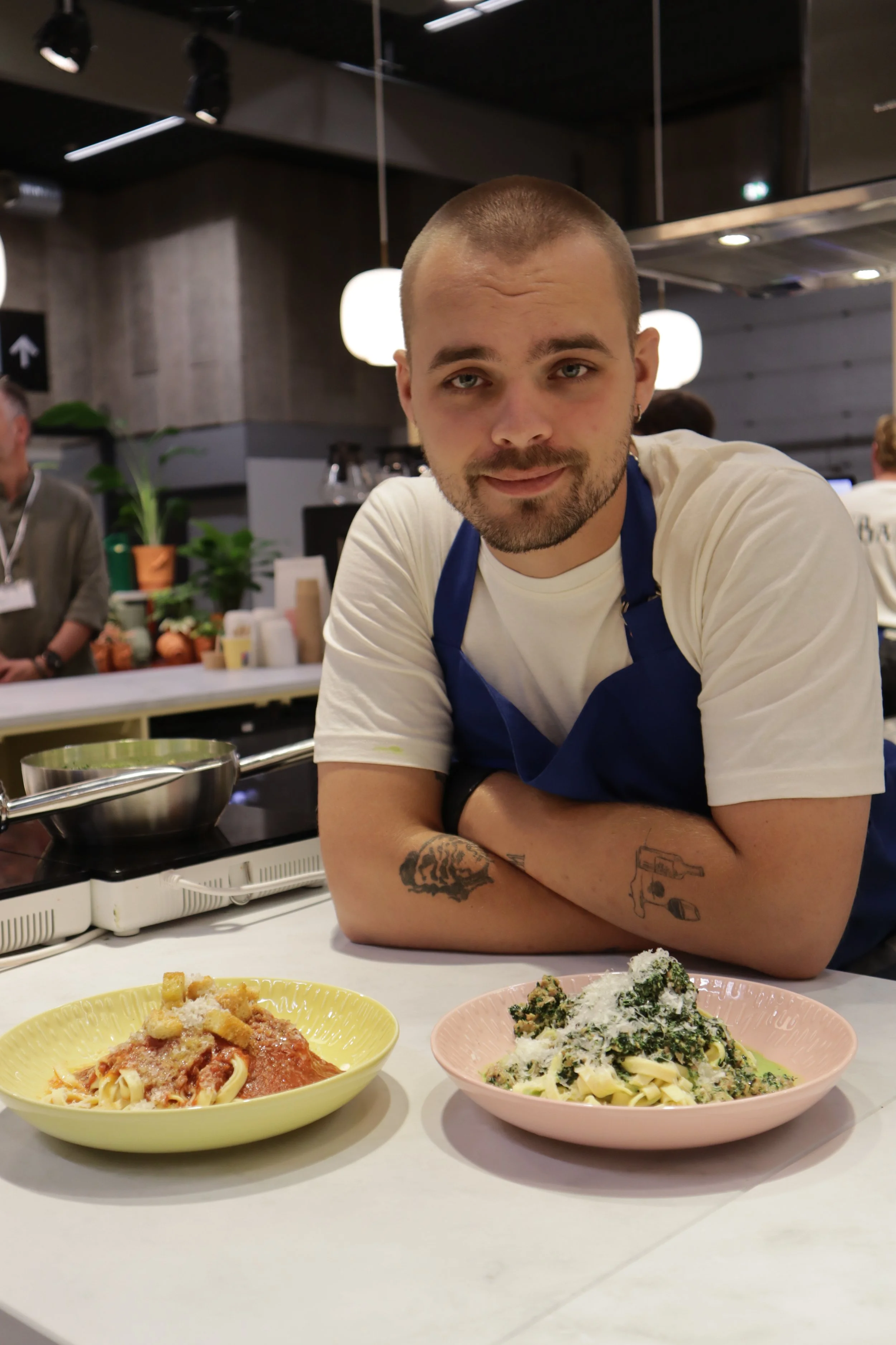 A man with tattoos on his forearms leaning on a kitchen counter with two plates of pasta in front of him, in a restaurant setting.