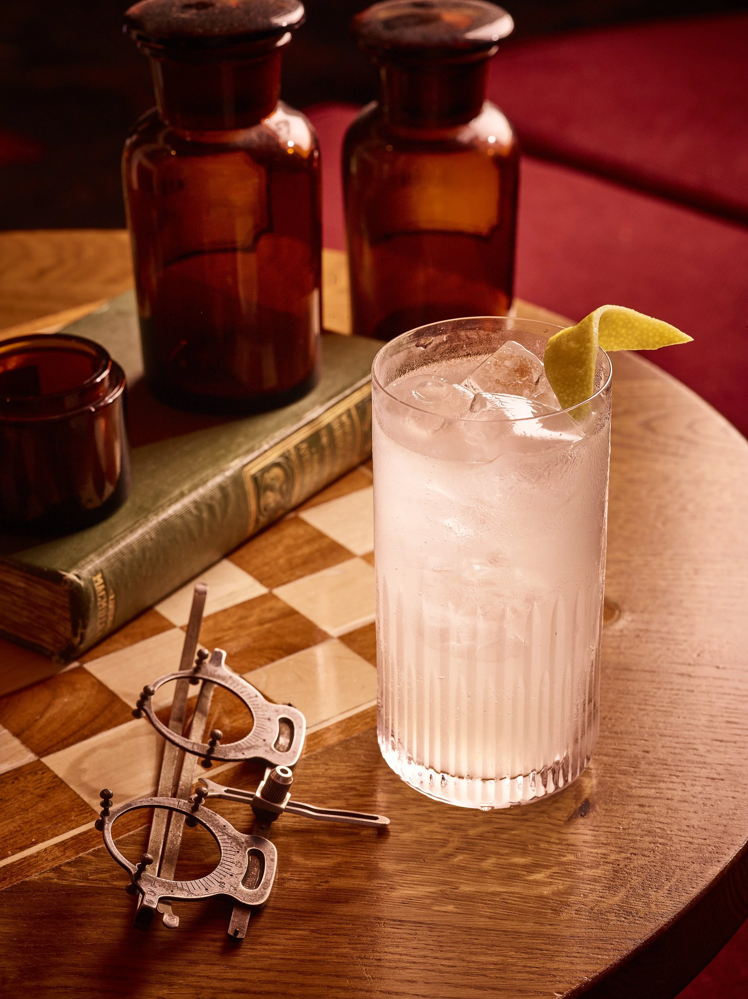 A wooden table with vintage brass tools, two amber glass bottles, and a clear glass of Wolfschmidts Kummel Cocktail recommendation - The kummel Spritz with a lemon twist and ice cubes.