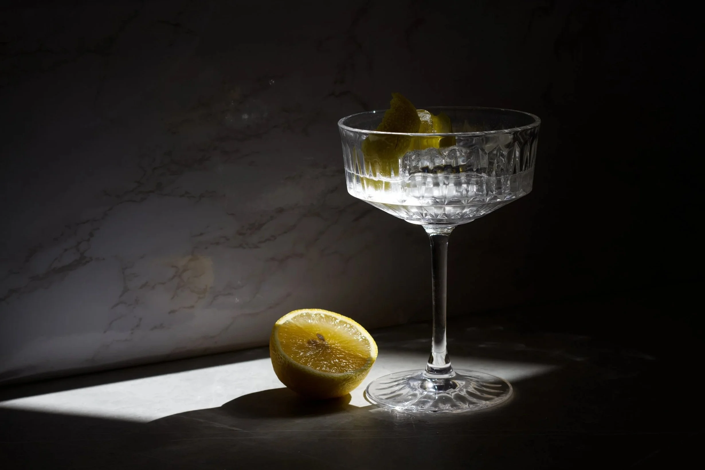 A clear glass of Wolfschmidt kümmels signature "Silver Bullet" cocktail, with a lemon twist inside, placed on a marble surface, with a lemon slice in front, illuminated against a dark background.