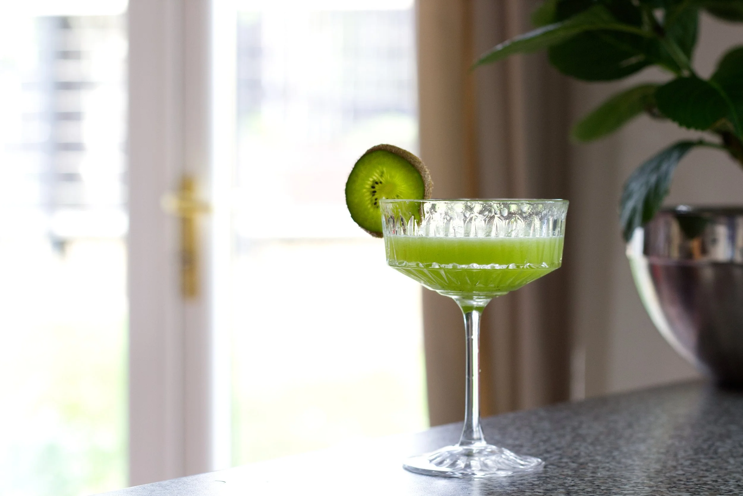 Wolfschmidt Kuumels Recommended Kummel Kiwi martini A glass filled with bright green kiwi juice, garnished with a slice of kiwi fruit on the rim, placed on a countertop with a window and green houseplant in the background.