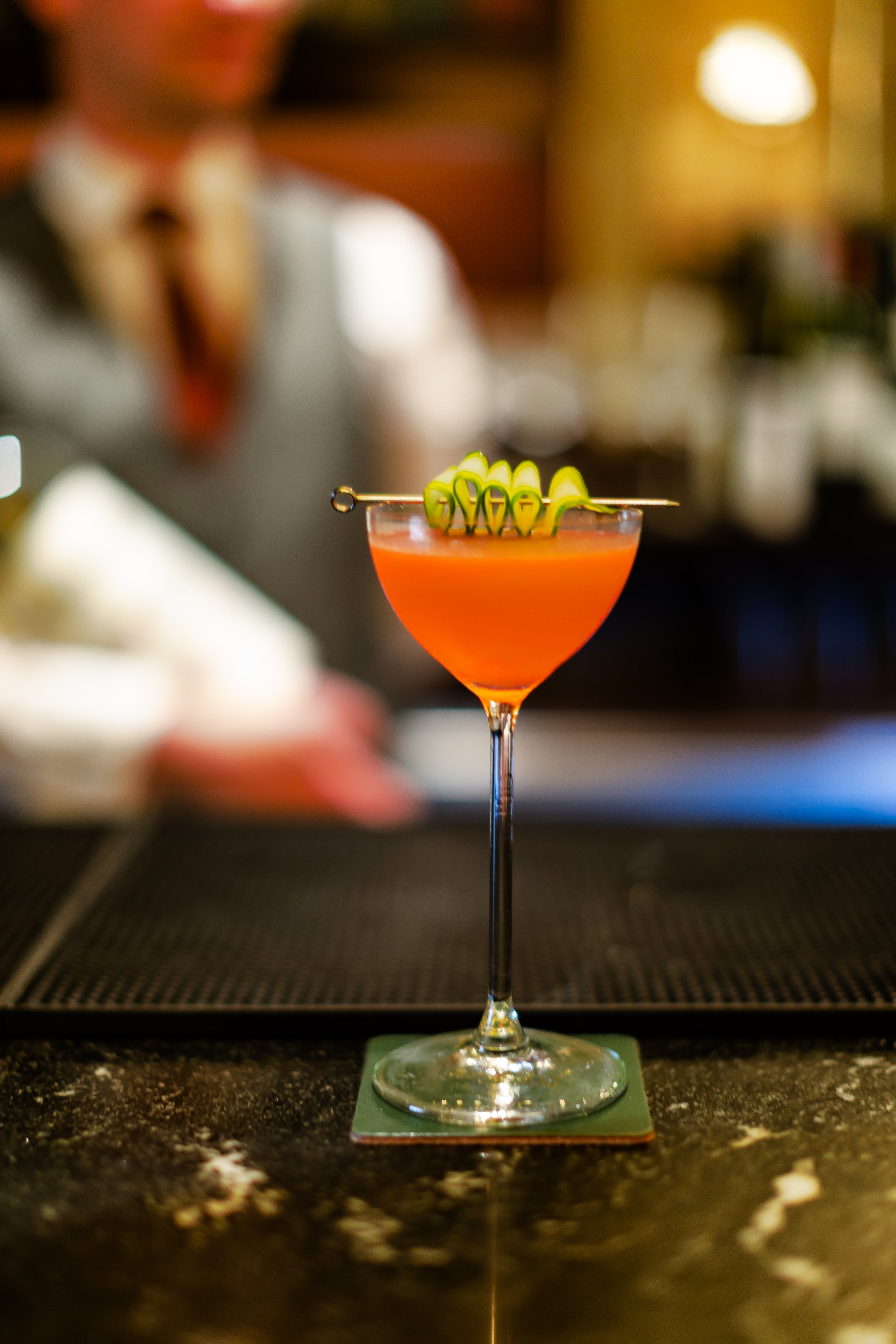The ribbon cocktail in a martini glass with a green garnish on top, on a bar counter, with a bartender in the background.
