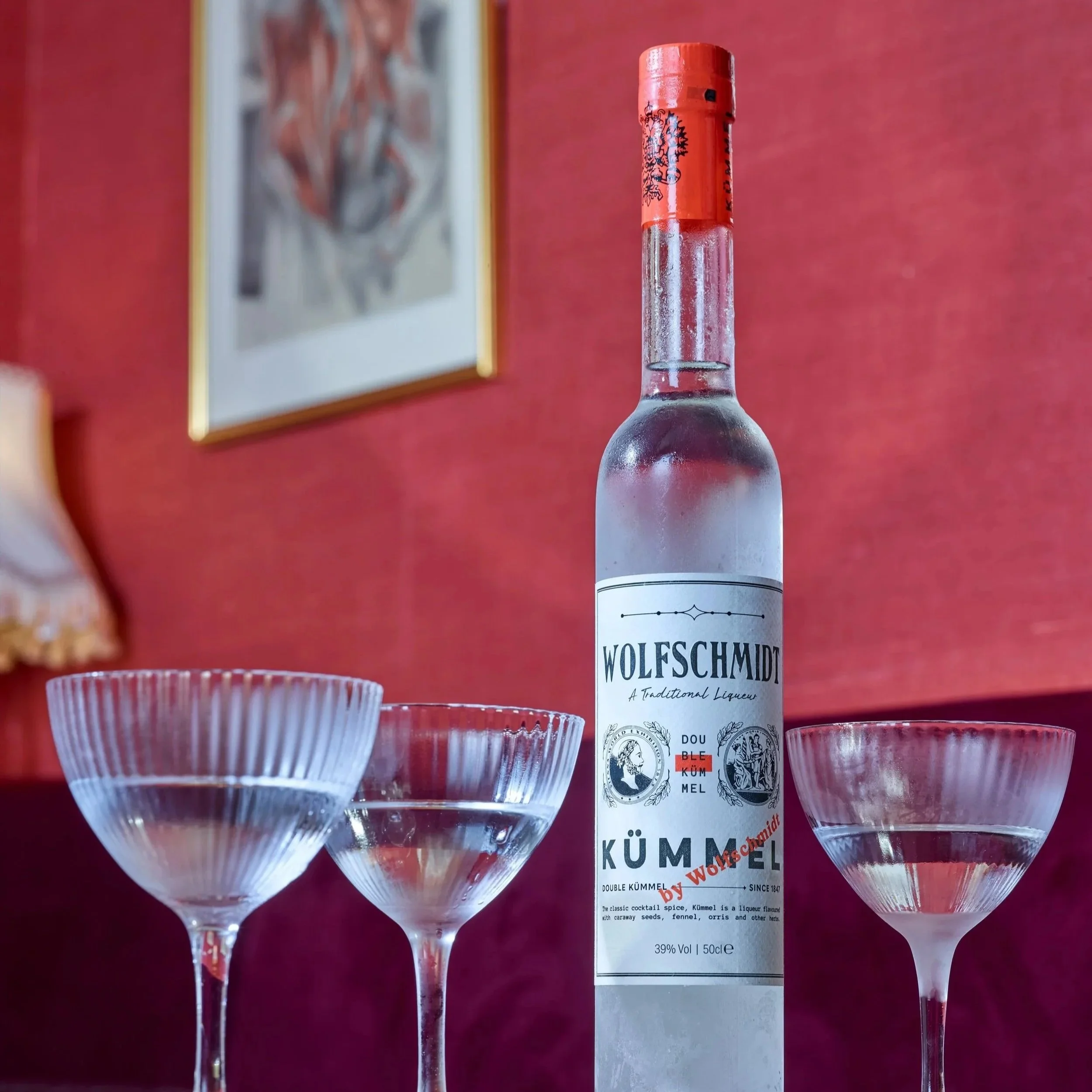 A clear glass bottle of Wolfschmidt Kümmel Luqueur surrounded by cocktail glasses on a table with a red wall and a framed abstract artwork in the background.