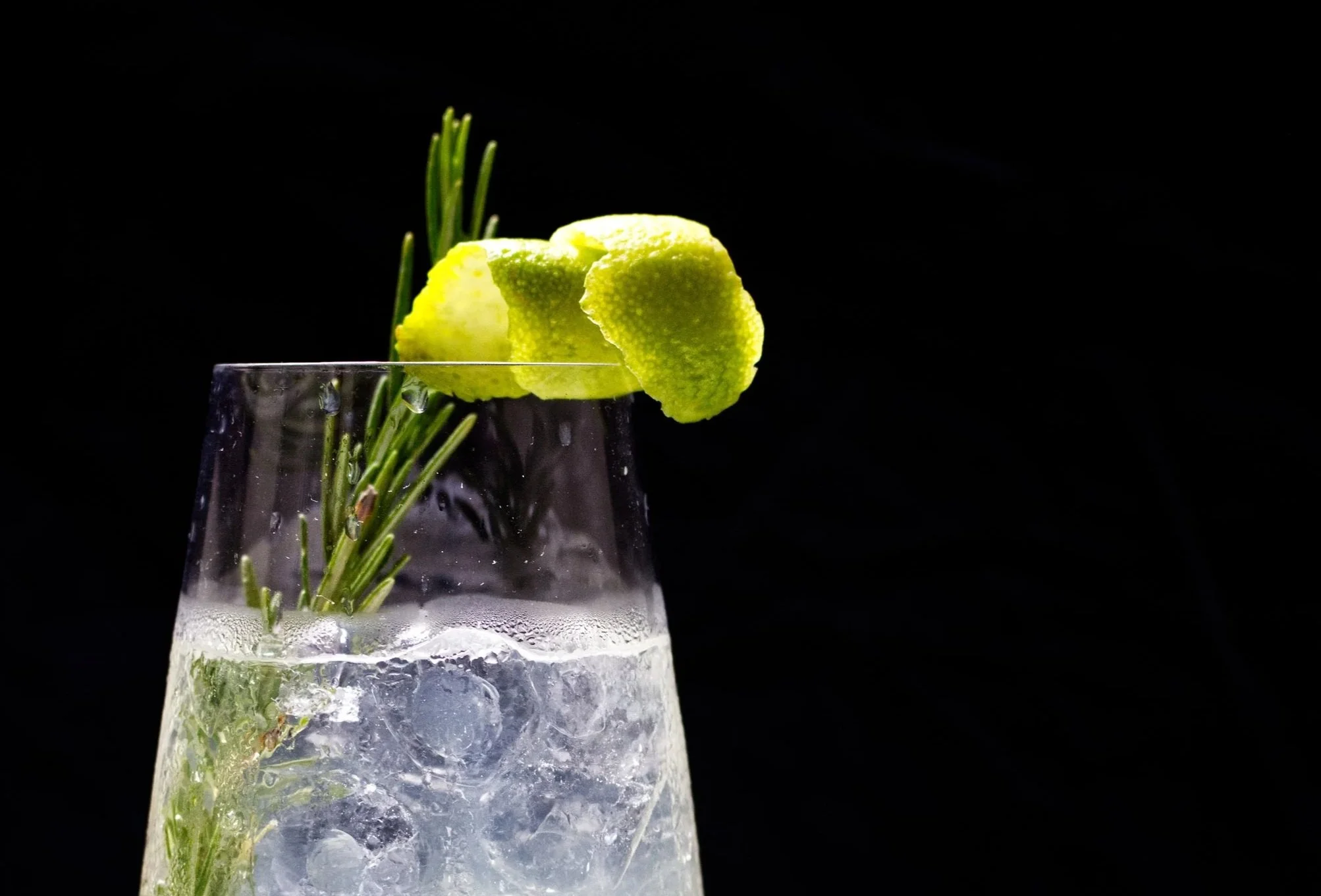 A clear Kummel Spritz glass contains ice, a sprig of rosemary, and lime slices, with a black background.