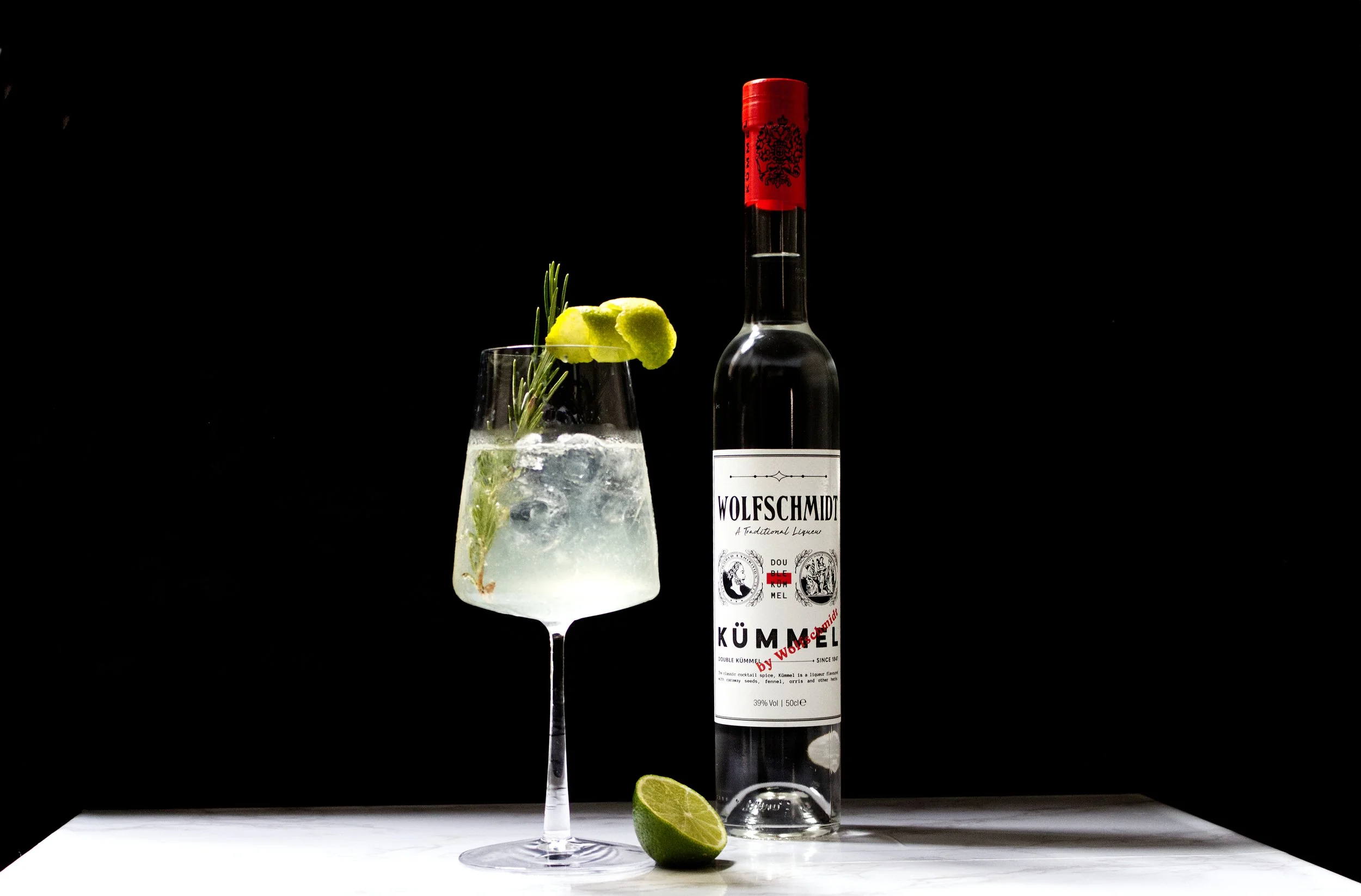 A tall Kümmel Spritz cocktail in a glass filled with ice, garnished with a lime wedge and a sprig of rosemary, with a bottle of Wolfschmidt Kümmel liqueur and a lime half placed on a white surface against a black background.