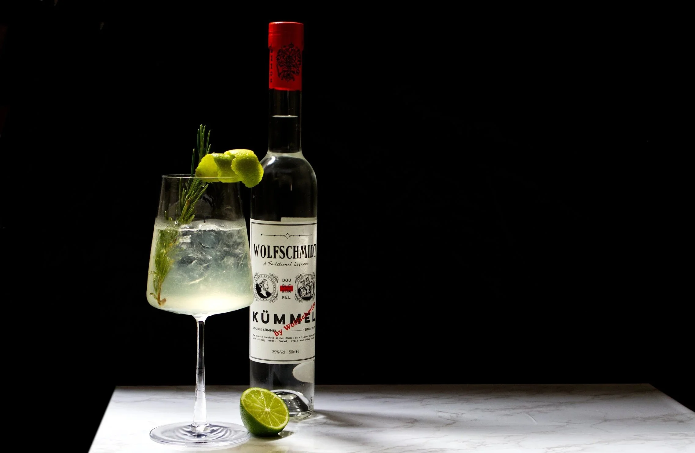 A Kummel cocktail glass filled with a light-colored drink, garnished with a sprig of herbs, lime slices, and ice, with a bottle of Wolfschmidt Kummel liqueur and a lime half on a white surface against a black background.