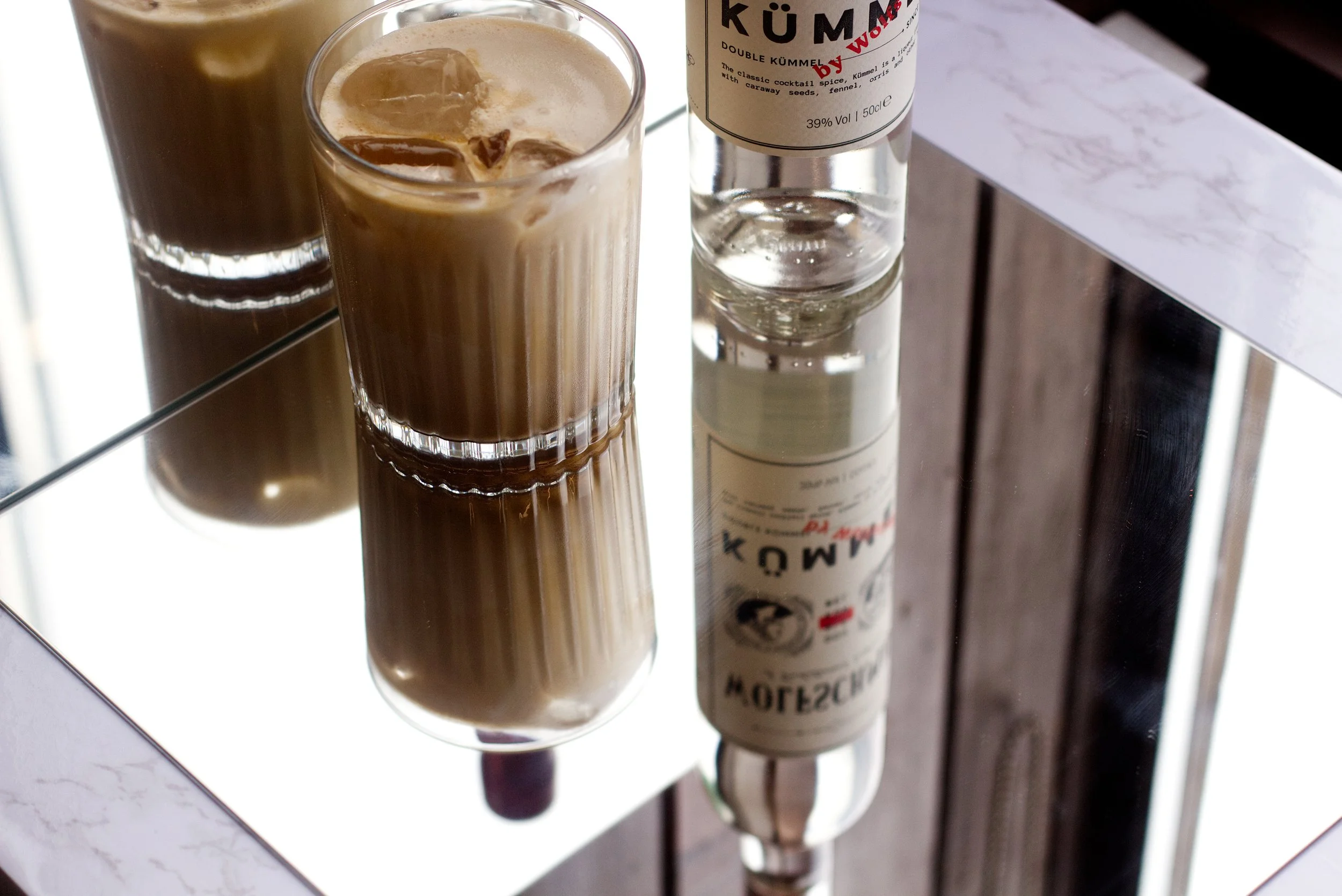 Wolfschmidt Kümmels recommended "After Hours" cocktail showing a creamy glass of iced coffee liqueur on a reflective surface, with a bottle of Kümmel liqueur nearby.